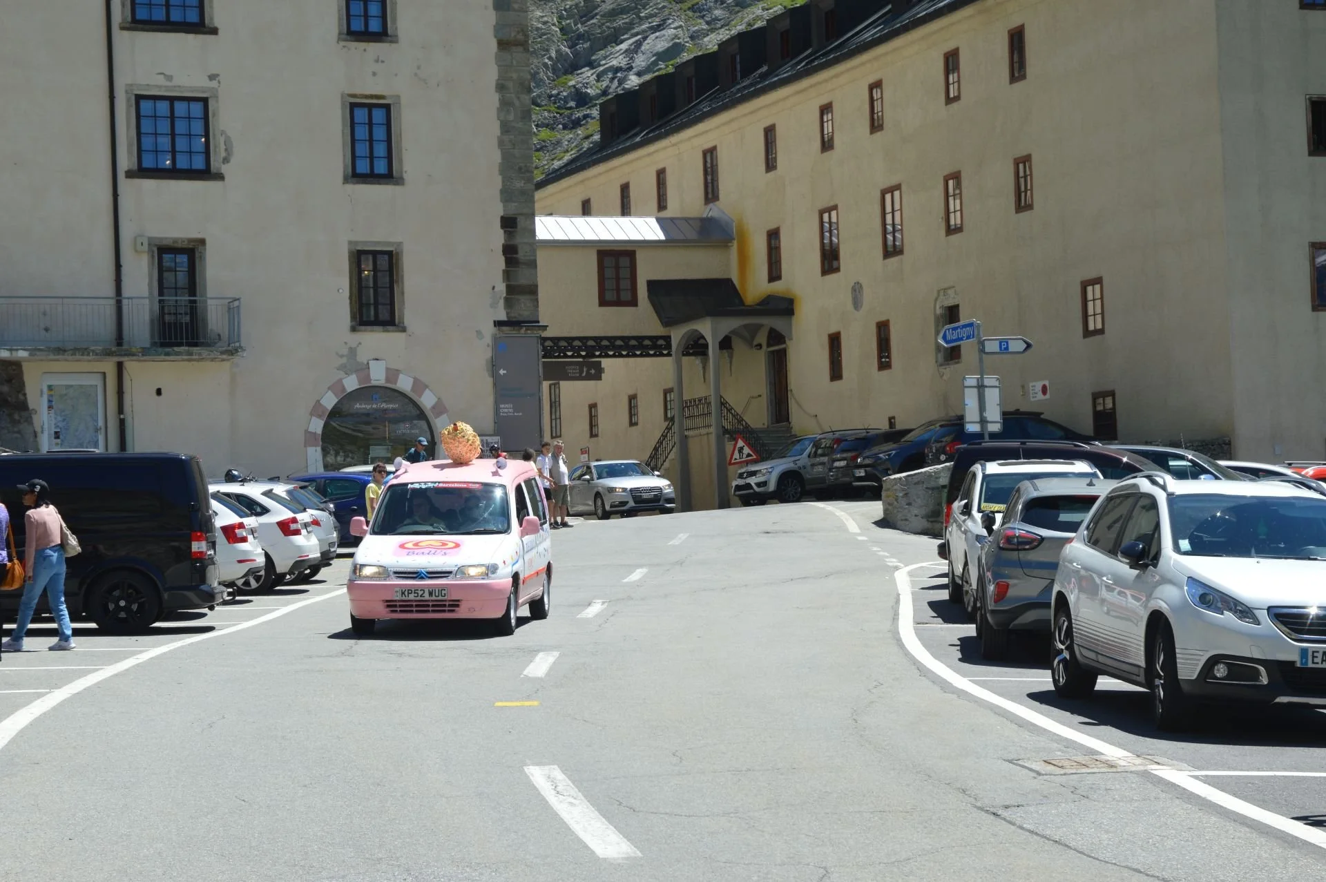 A parking lot with several parked cars and a pink van with an ice cream cone on top, situated near beige buildings with multiple windows. Pedestrians are walking along the area and there are street signs, including a blue one pointing to Martigny.