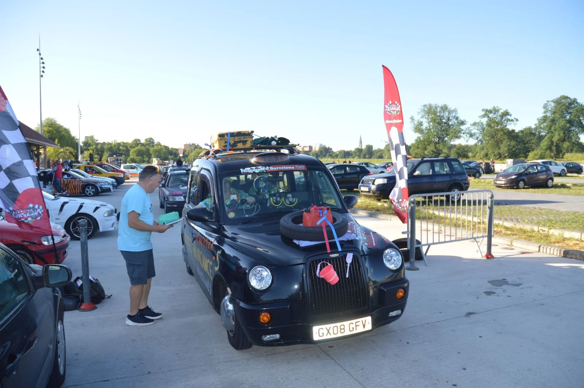 A black car decorated with racing and automotive accessories at a car event, with a man reading a booklet nearby, surrounded by other parked cars and flags in a parking lot during daytime.