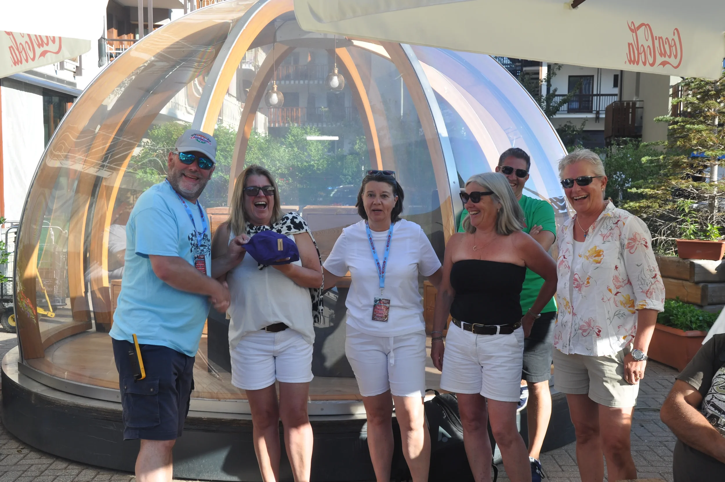 Six adults, four women and two men, stand together outdoors in front of a transparent dome structure, smiling and laughing.