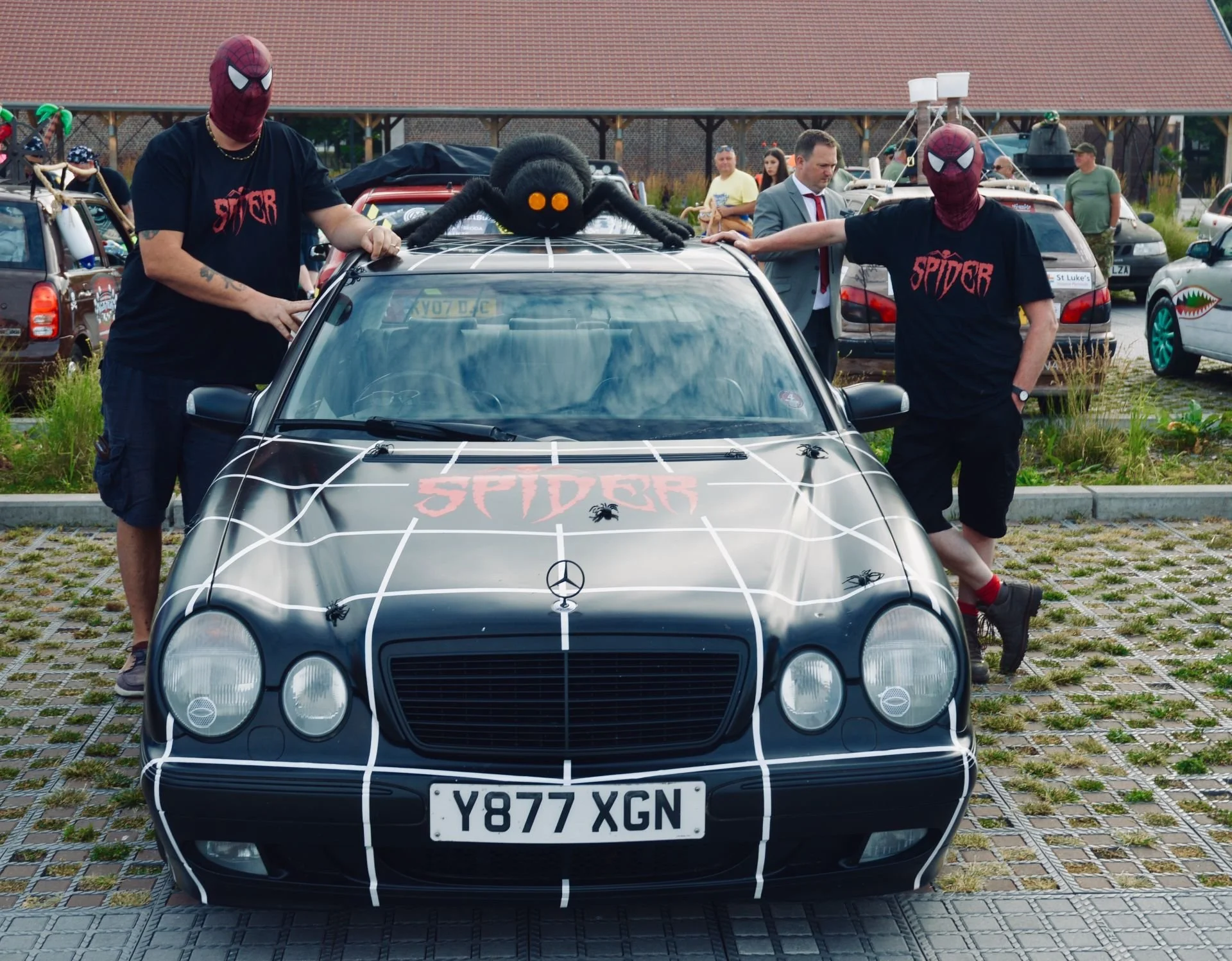 A black Mercedes-Benz car decorated with white spiderweb designs and spiders, with a red 'Spider' logo on the hood. Two men in Spider-Man masks and black shirts with 'Spider' written on them stand beside the car. A large plush spider sits on the roof