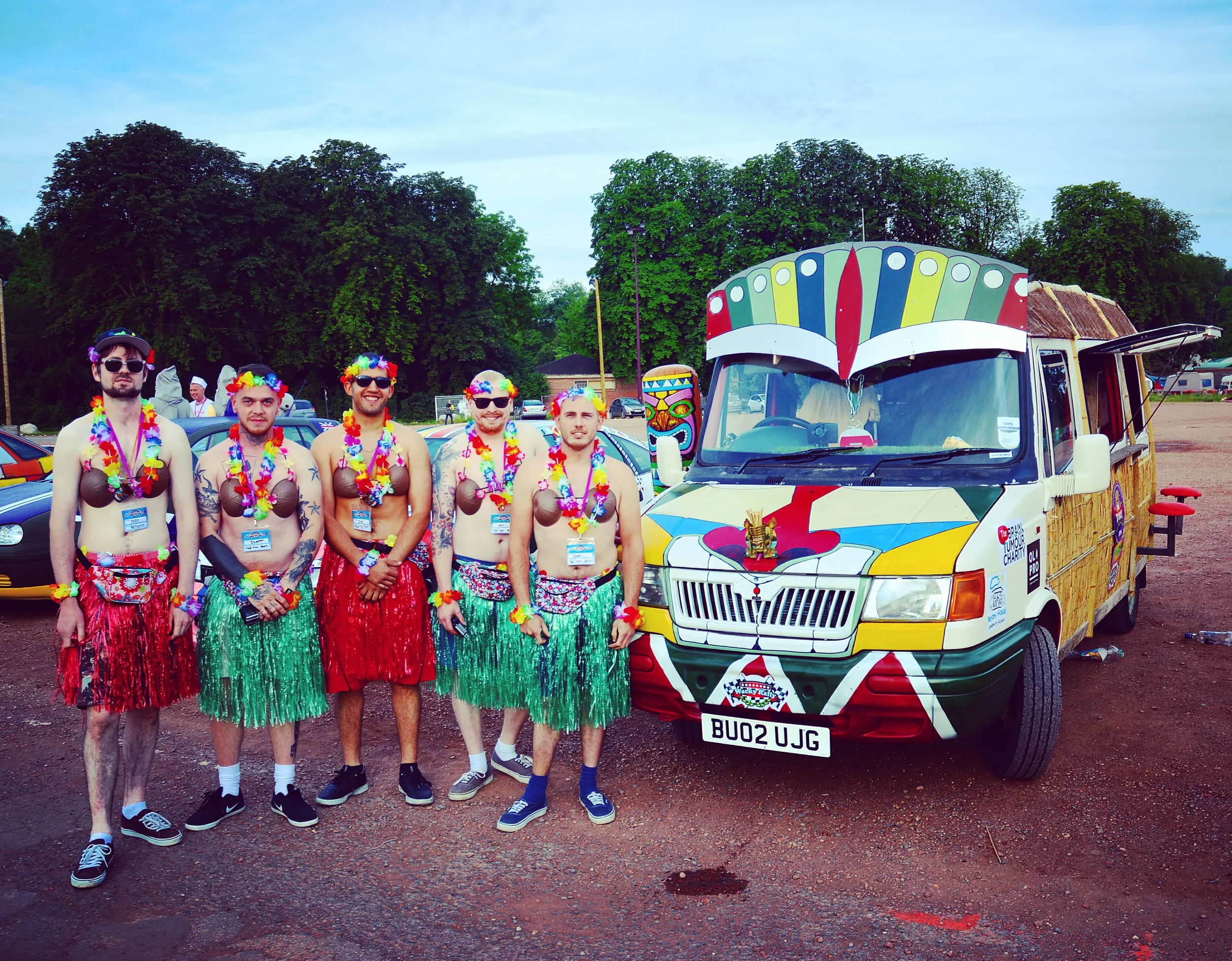 Group of five men dressed in colorful costumes with leis, grass skirts, and sunglasses standing next to a decorated van at an outdoor event with trees in the background.