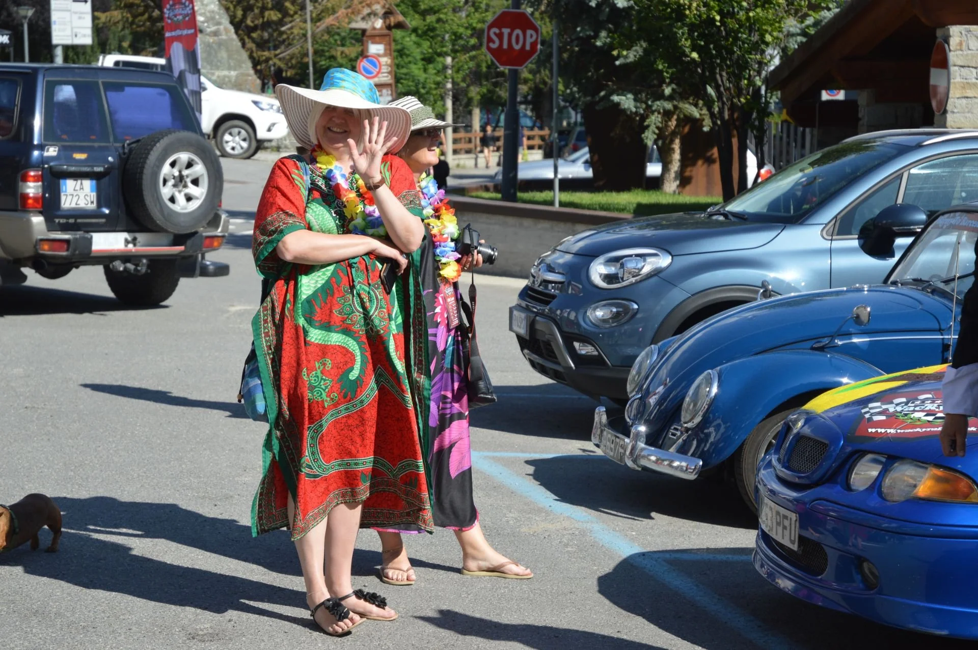 Two women dressed in colorful, tropical-themed clothing with leis around their necks, wearing large sun hats, standing in a parking lot, smiling and waving at the camera. There are several cars parked nearby and a stop sign in the background.
