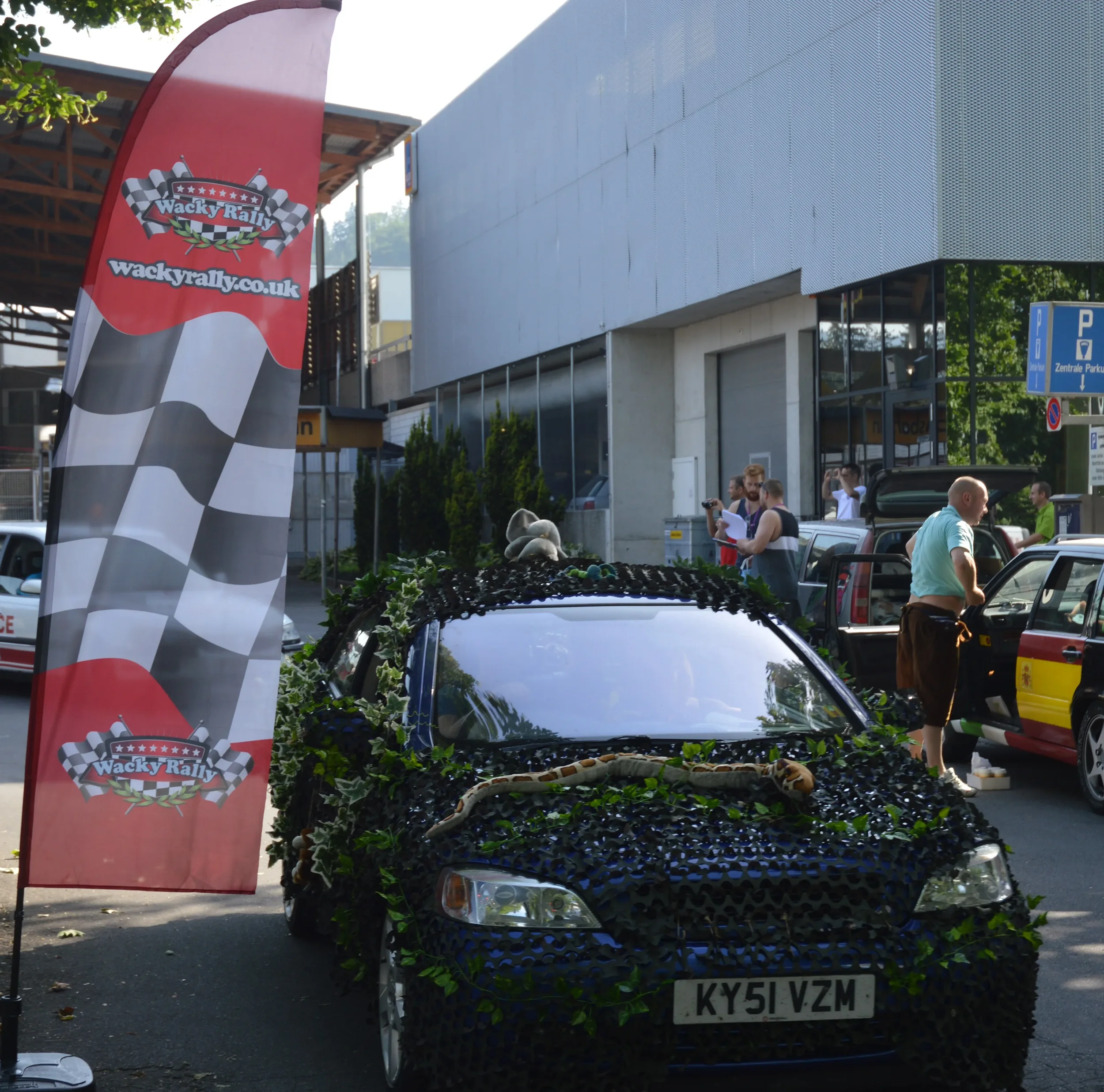 Car decorated with fake plants and a snake lying on its front hood, parked near a Wacky Rally event banner, with several people standing and talking in the background.