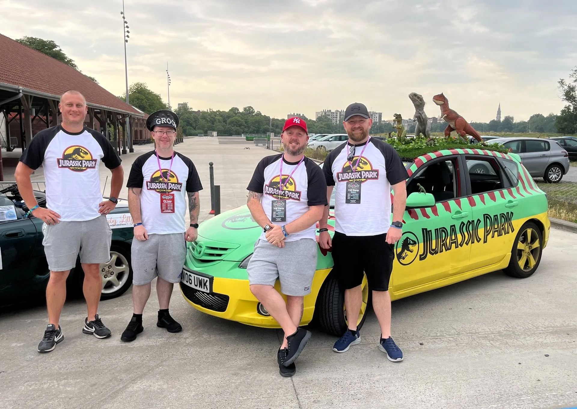 Four men standing in front of a green and yellow Jurassic Park-themed car, all wearing Jurassic Park t-shirts and badges, with Dino sculptures on top of the car, in a parking lot.