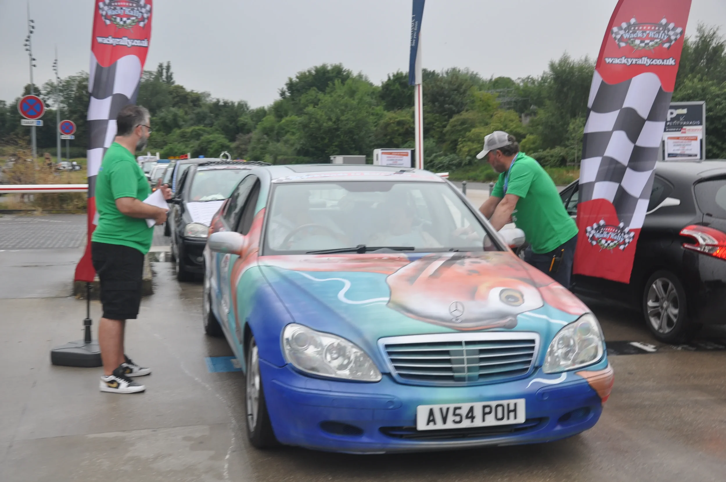 A Mercedes-Benz car with a colorful turtle painted on the hood, parked at a rally event with two men in green shirts beside it, and race flags in the background.