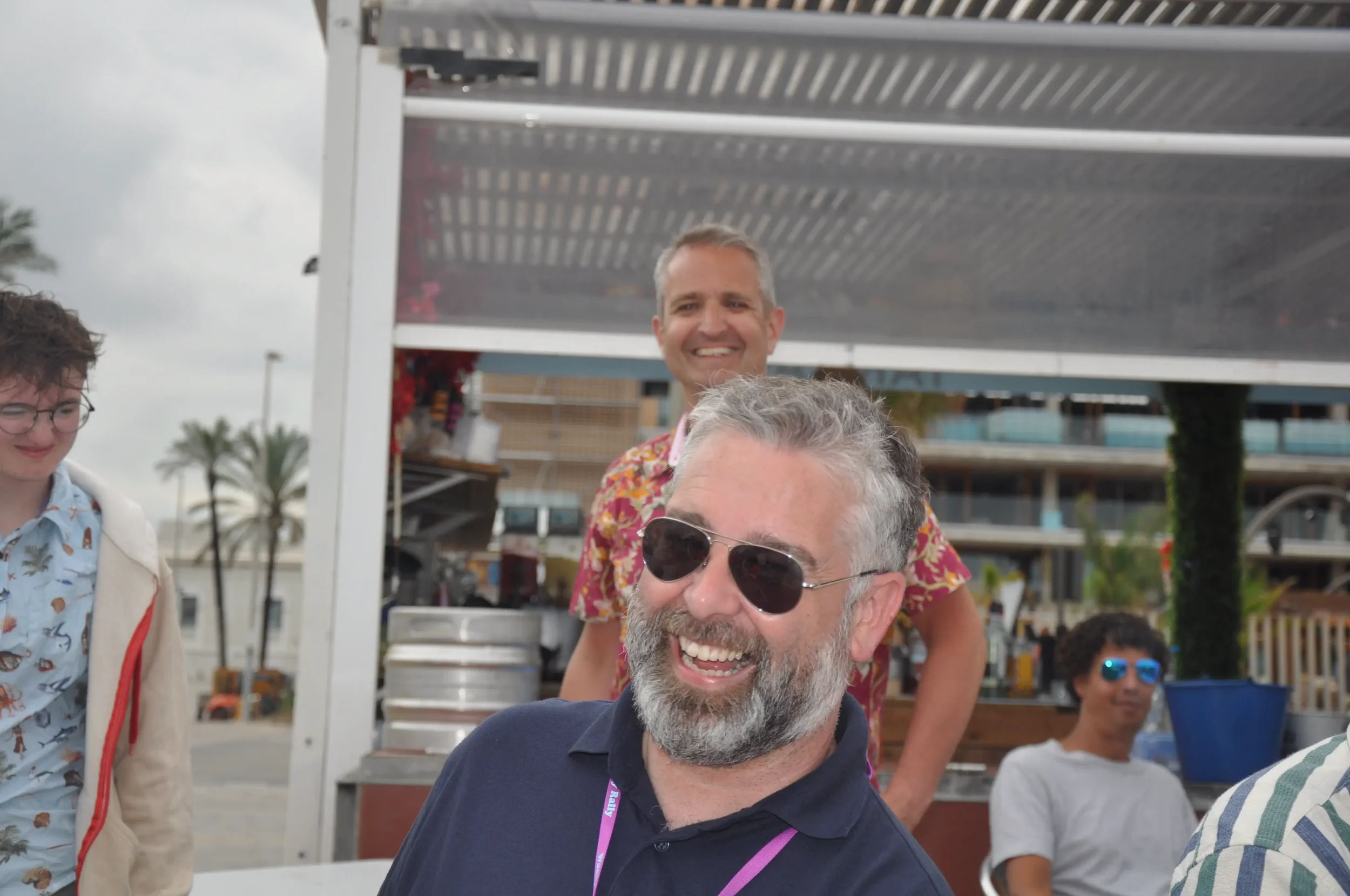 Group of people outdoors enjoying a sunny day, smiling, with a man wearing sunglasses and a navy polo in the foreground. Other individuals are in the background near a food stand with a cityscape and palm trees visible.