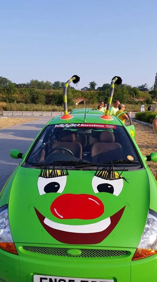 A bright green car decorated as a cartoon clown face with large eyes, a red nose, and a big smile, parked outdoors with people nearby, some wearing yellow shirts, and two large striped antennas on the roof.