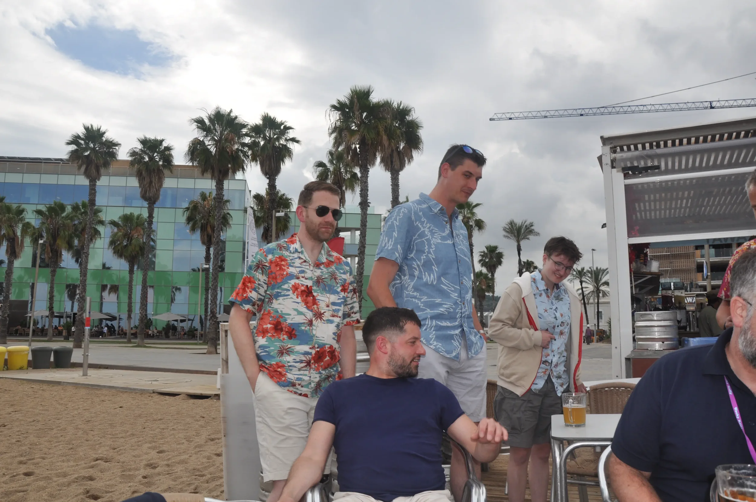 Group of five men at an outdoor beachside venue, with palm trees, sandy ground, and a modern glass building in the background. Two men are sitting at a table, and three are standing behind them. The men are wearing casual, summer-style clothing.