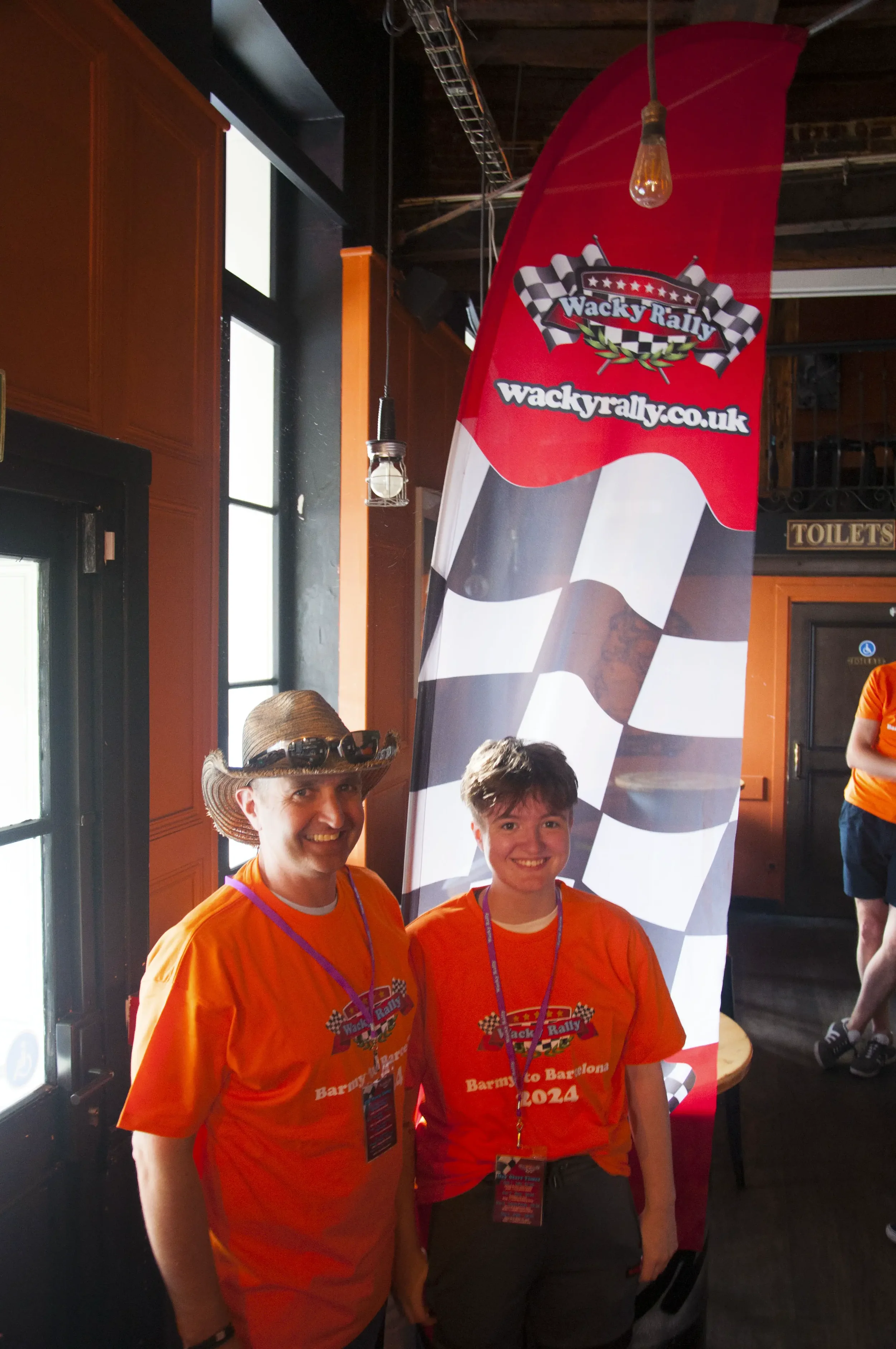 Two smiling individuals wearing bright orange T-shirts with a rally logo, standing inside a venue near a race flag and a tall banner that reads "Wacky Rally" with a checkered flag pattern and website URL, participating in a rally event.