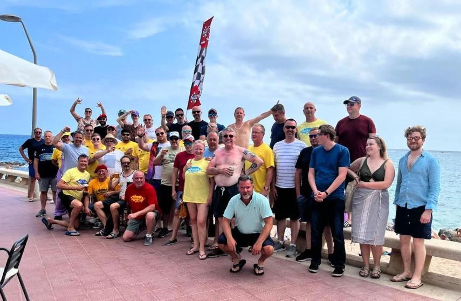 Group of people gathered on a seaside promenade, some in swimsuits, with an ocean and a cloudy sky in the background, celebrating or socializing.