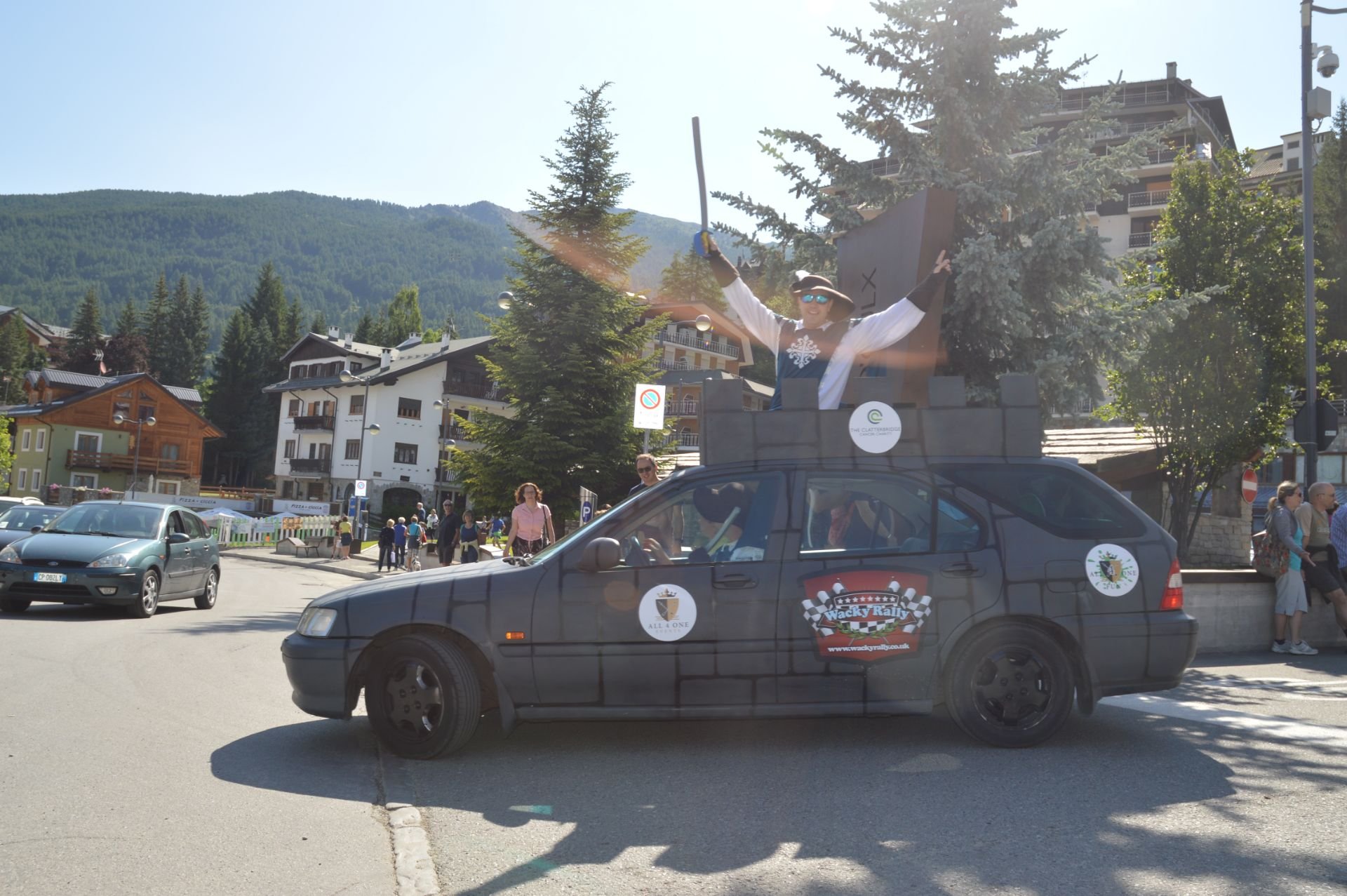 A person dressed as a medieval knight standing in the back of a decorated black vehicle with a castle wall design, holding a sword and raising one arm, at a parade or festival in a village square with trees, mountains, and spectators in the backgroun