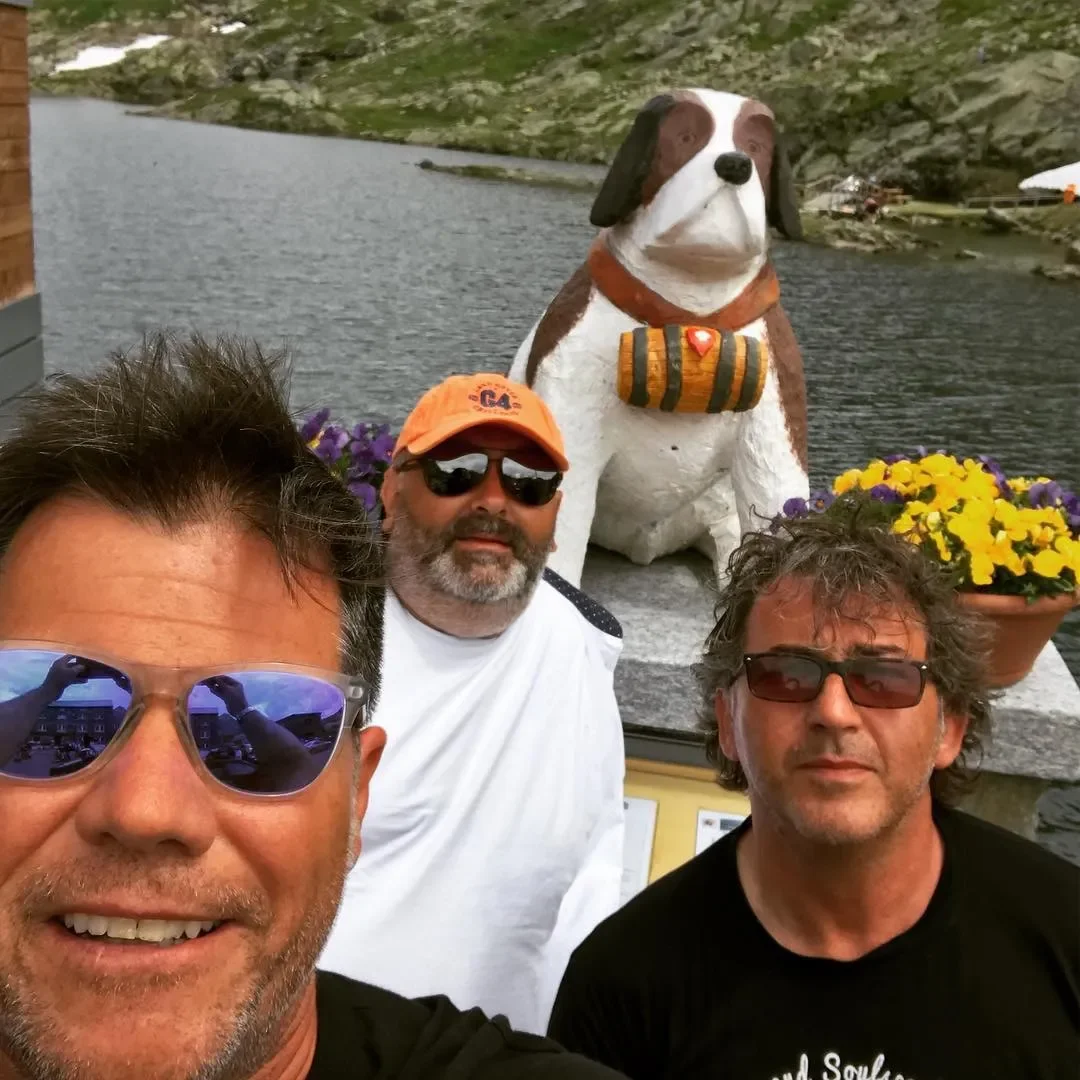 Three men taking a selfie in front of a large dog sculpture with a lake and mountains in the background and purple and yellow flowers in pots nearby.
