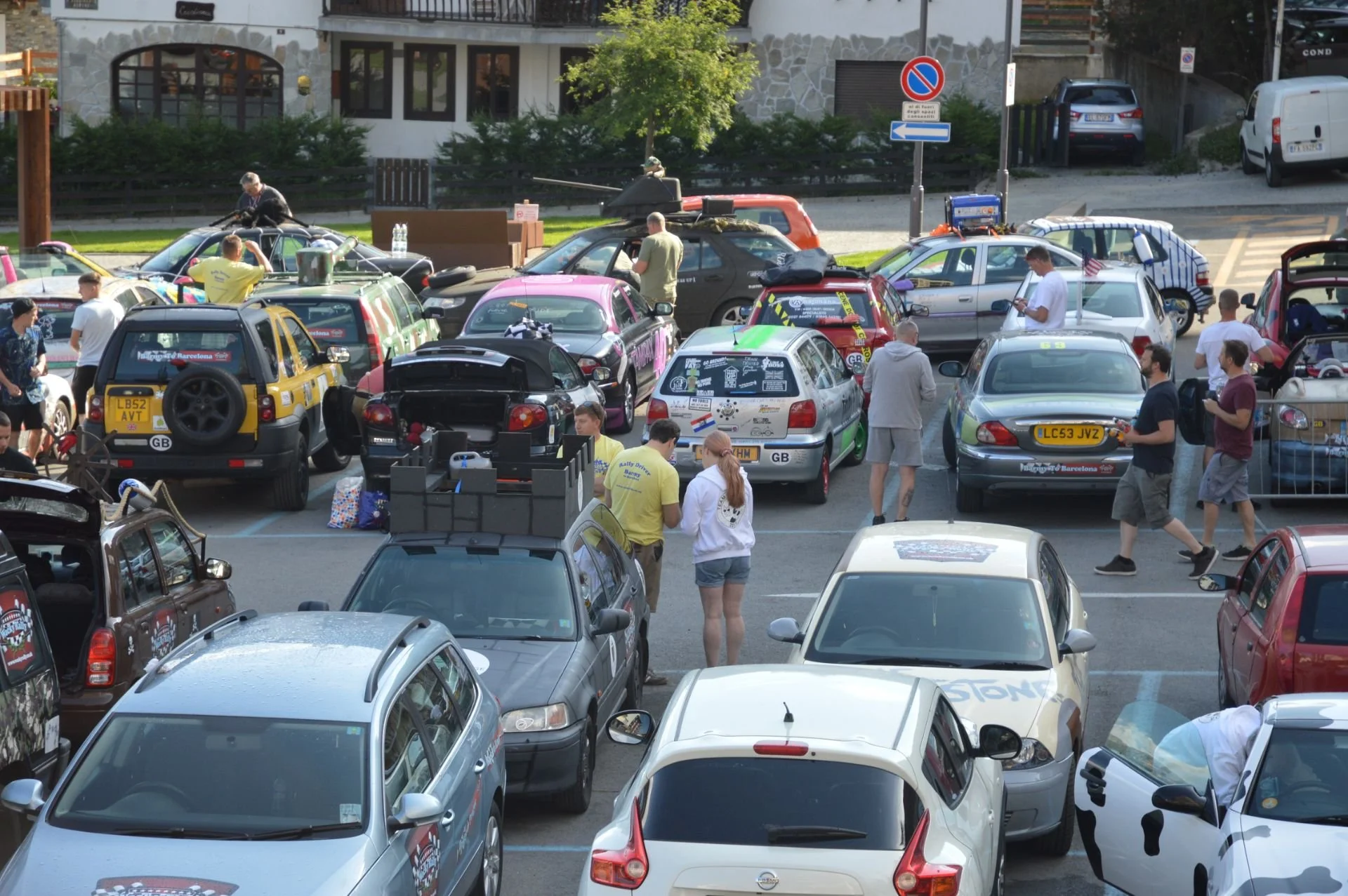 A busy parking lot filled with various cars and people. Some cars have stickers and markings, suggesting a car event or rally. People are walking, chatting, and inspecting vehicles, with some engaged in activities around the cars. The background show