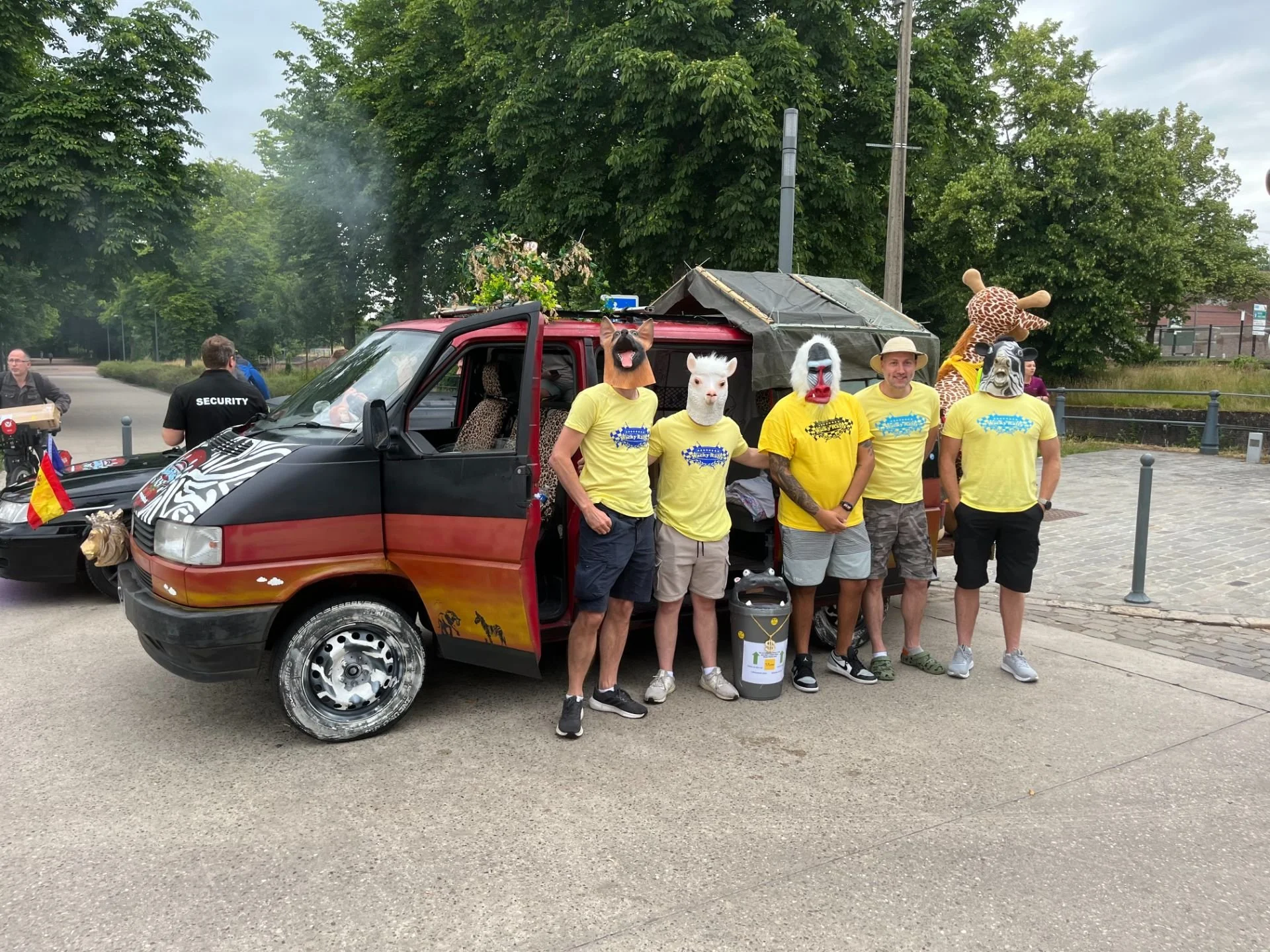 Group of five people wearing yellow shirts and animal masks, standing in front of a decorated small van with giraffe plush, in an outdoor area with trees and cloudy sky.
