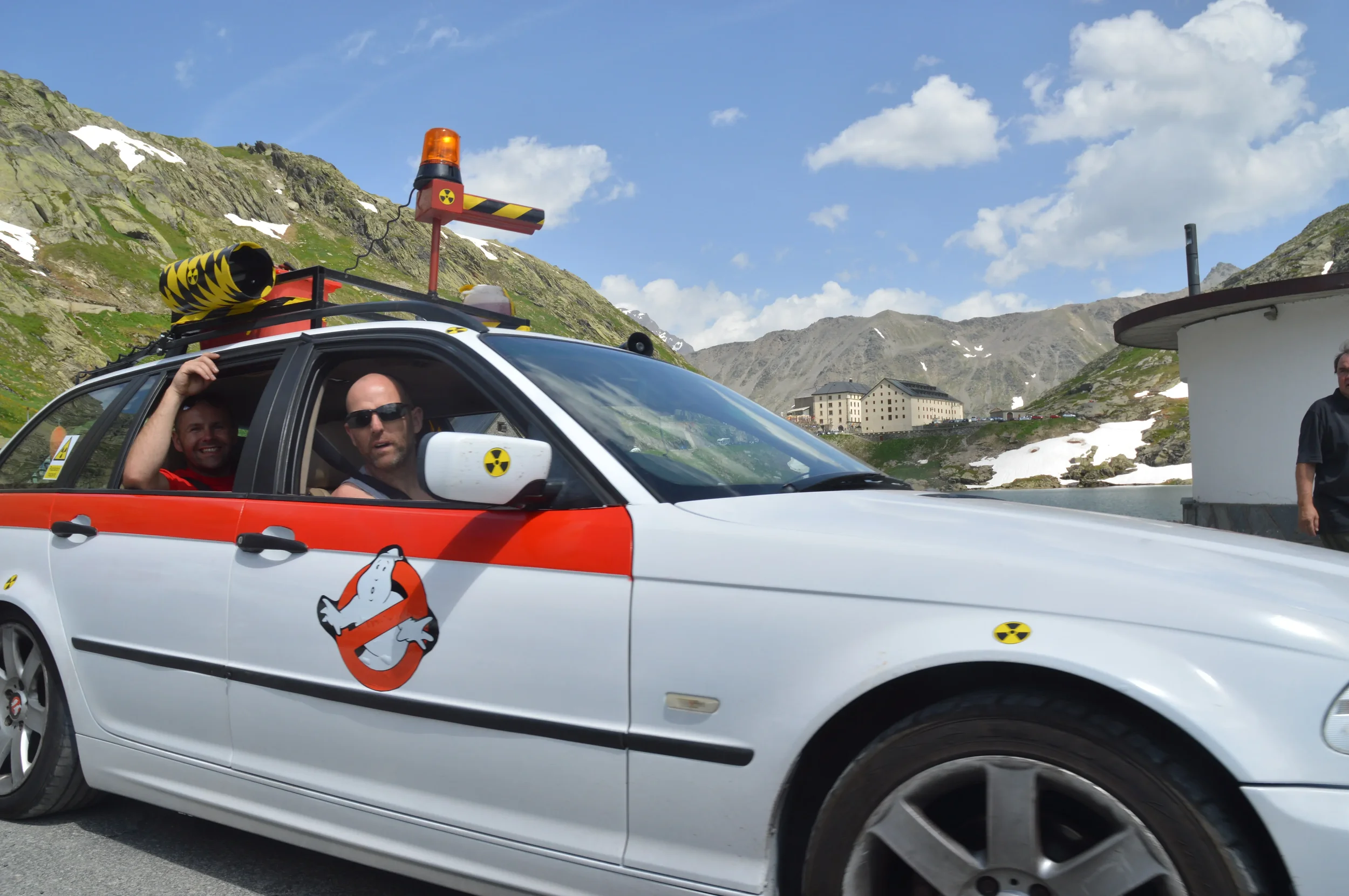 A white police car with Ghostbusters logo on the door, equipped for hazardous conditions, with two men inside, one wearing sunglasses and the other smiling. The car is parked on a mountain road with snowy patches, rocky terrain, and a large building 