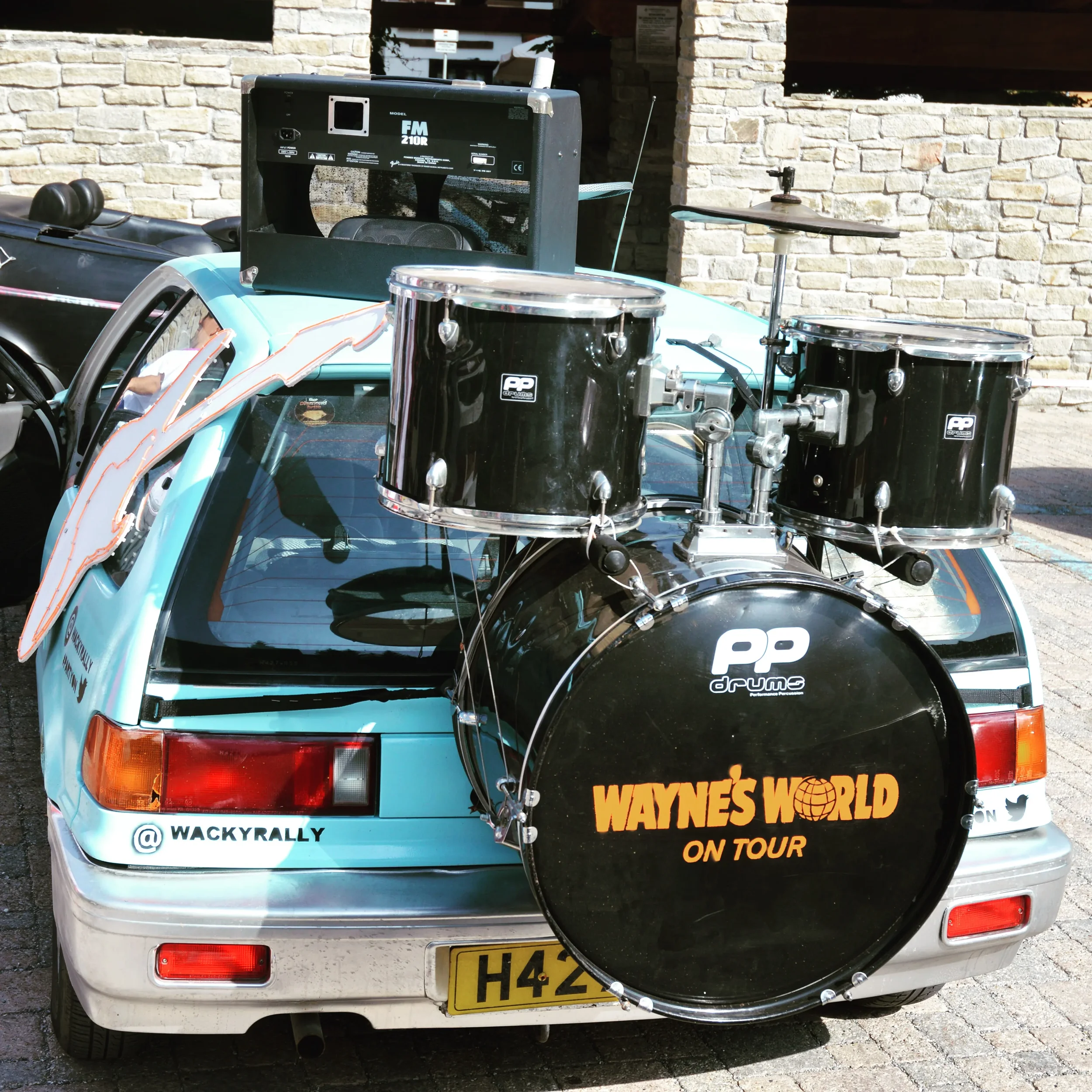 Rear view of a light blue car with a drum set mounted on the back, including a bass drum with 'Wayne's World on Tour' written on it, and a small electronic music device on top of the car.