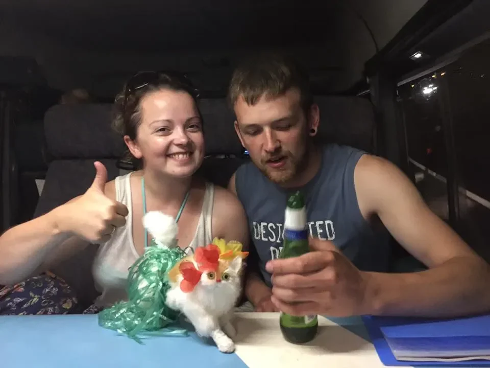 Two young adults sitting inside a vehicle. The woman on the left is smiling and giving a thumbs up, while the man on the right is looking at a bottle in his hand. There is a small decorative cat figure with flowers on its head between them. The woman