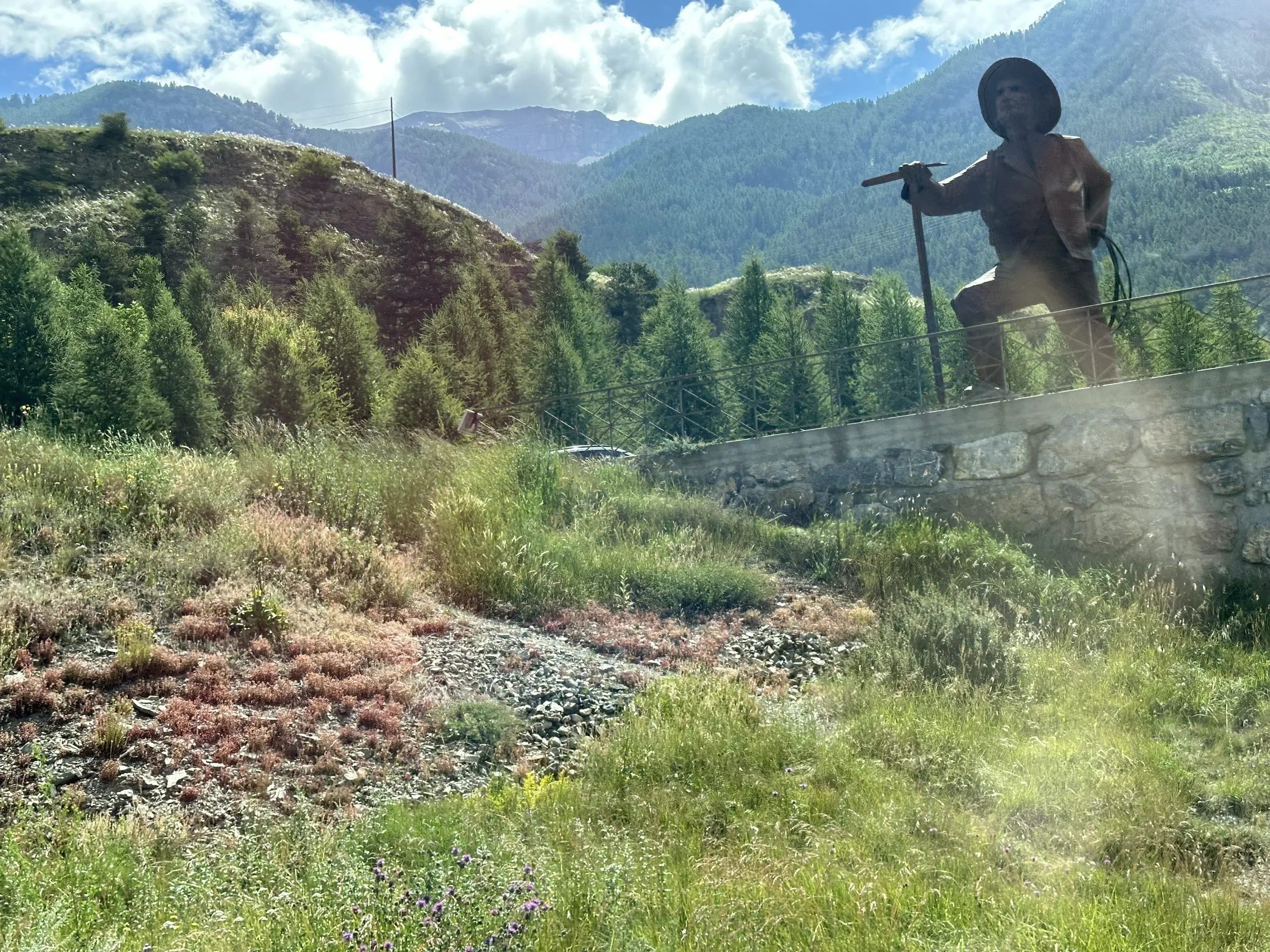 A large bronze statue of a man with a hat, holding a cane, seated on a stone wall in a lush, mountainous landscape with trees and hills in the background.