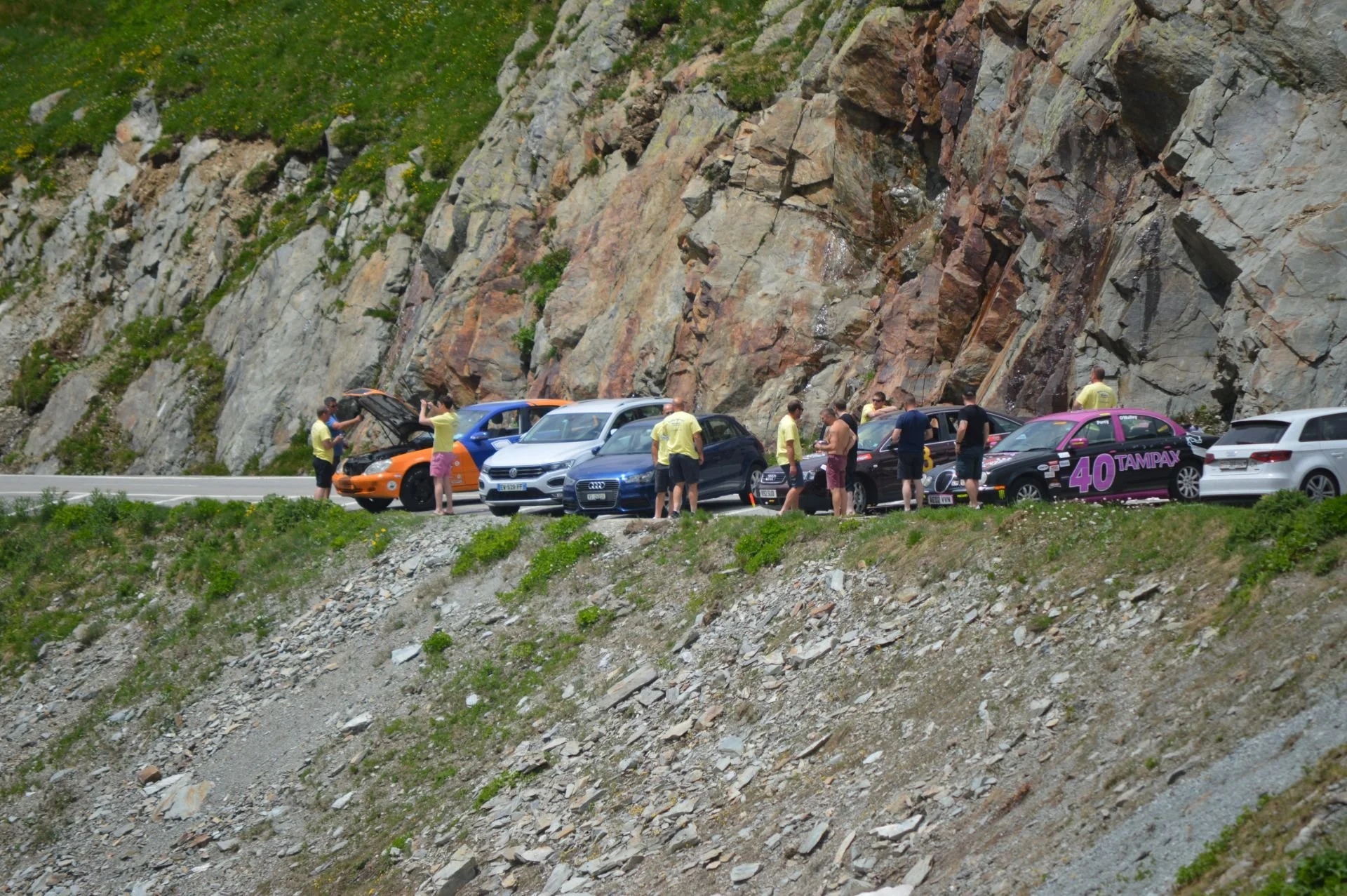 People standing around cars parked on a mountain road with rocky cliffs and greenery in the background.