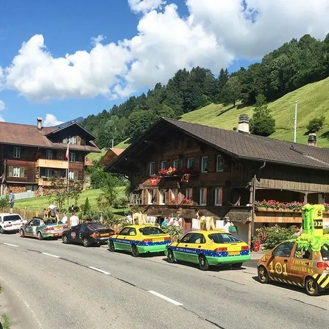 A mountain village street with several parked cars, including a yellow and green police car, a gold car with advertising, and others. There are people on the sidewalk, a large wooden chalet-style building with flower boxes on the windows, and green h