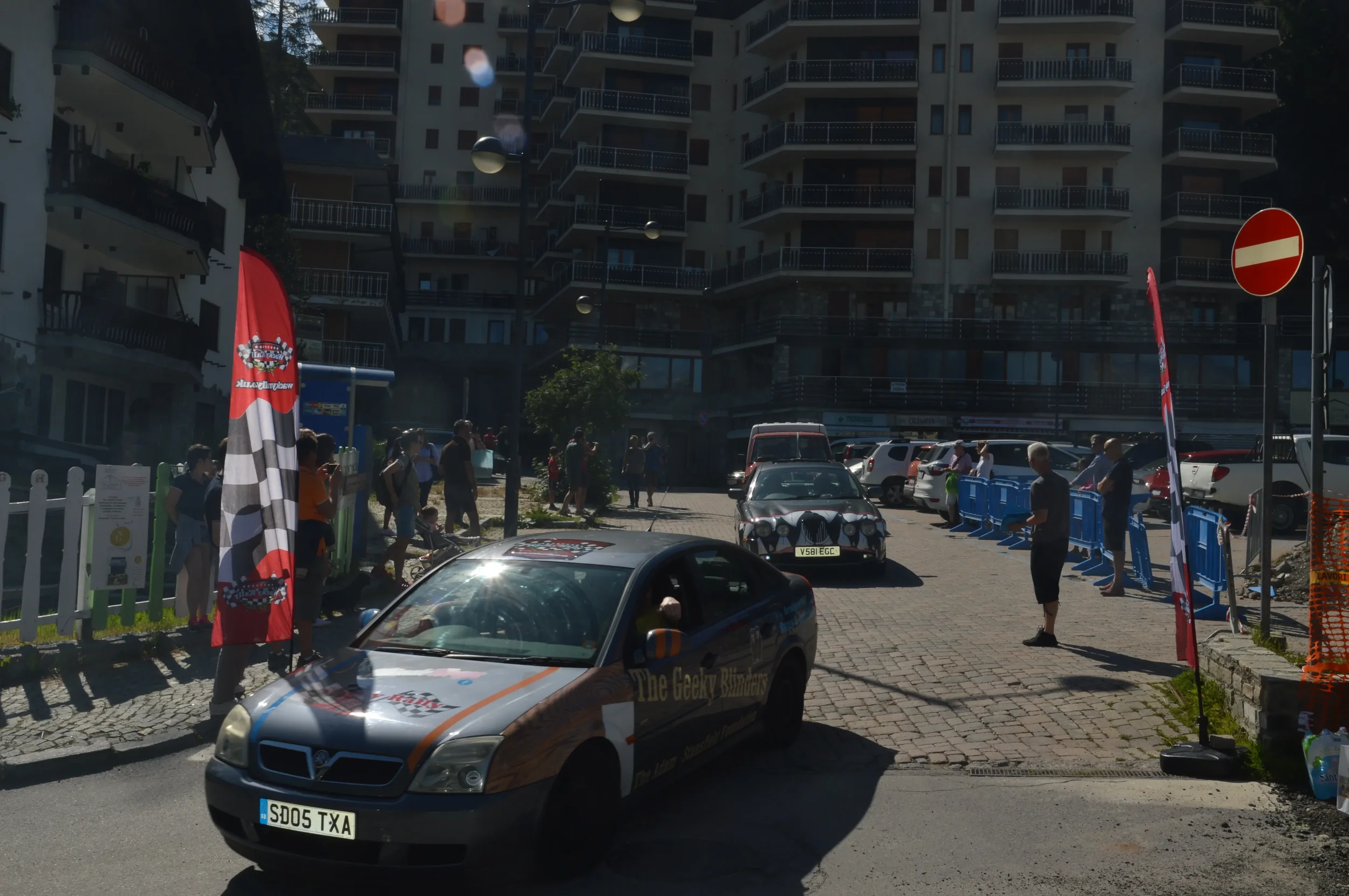 Street scene with cars, people and banners, residential buildings in the background, and a no entry sign.