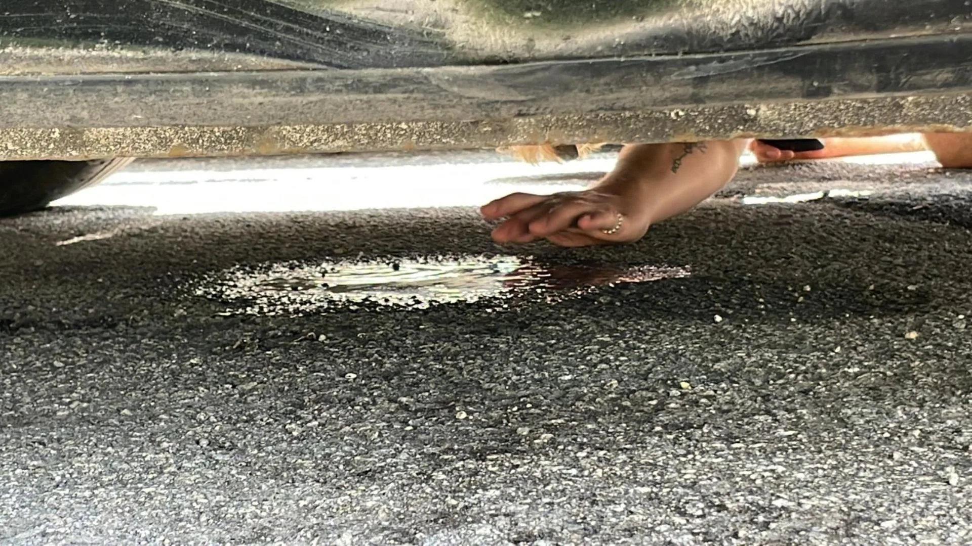 Person with a tattoo on their arm and multiple rings on their fingers taking a picture of a puddle under a car on asphalt pavement.