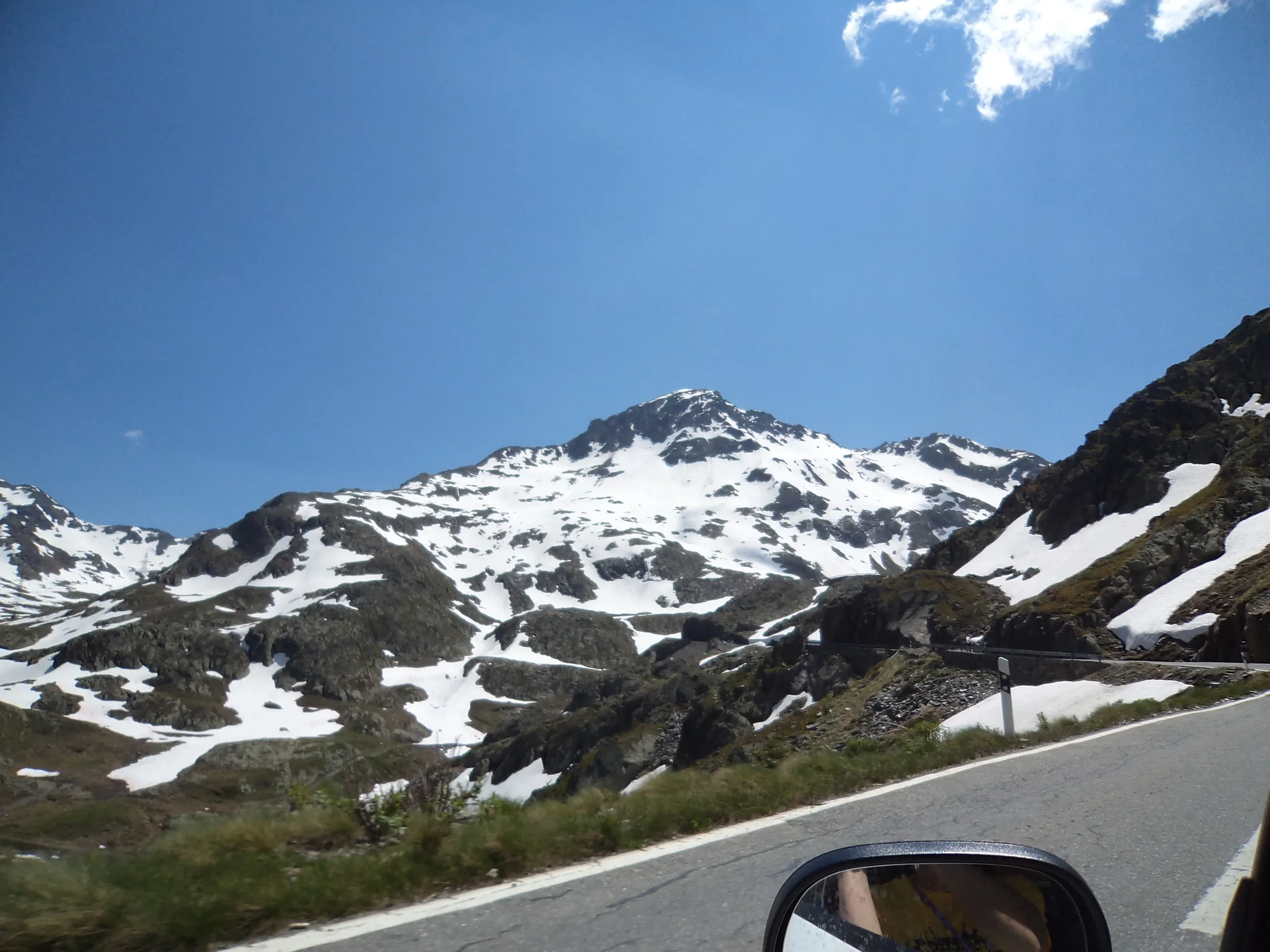 Snow-covered mountain landscape with rocky peaks, a clear blue sky, and a mountain road with a guardrail and a side mirror visible in the foreground.