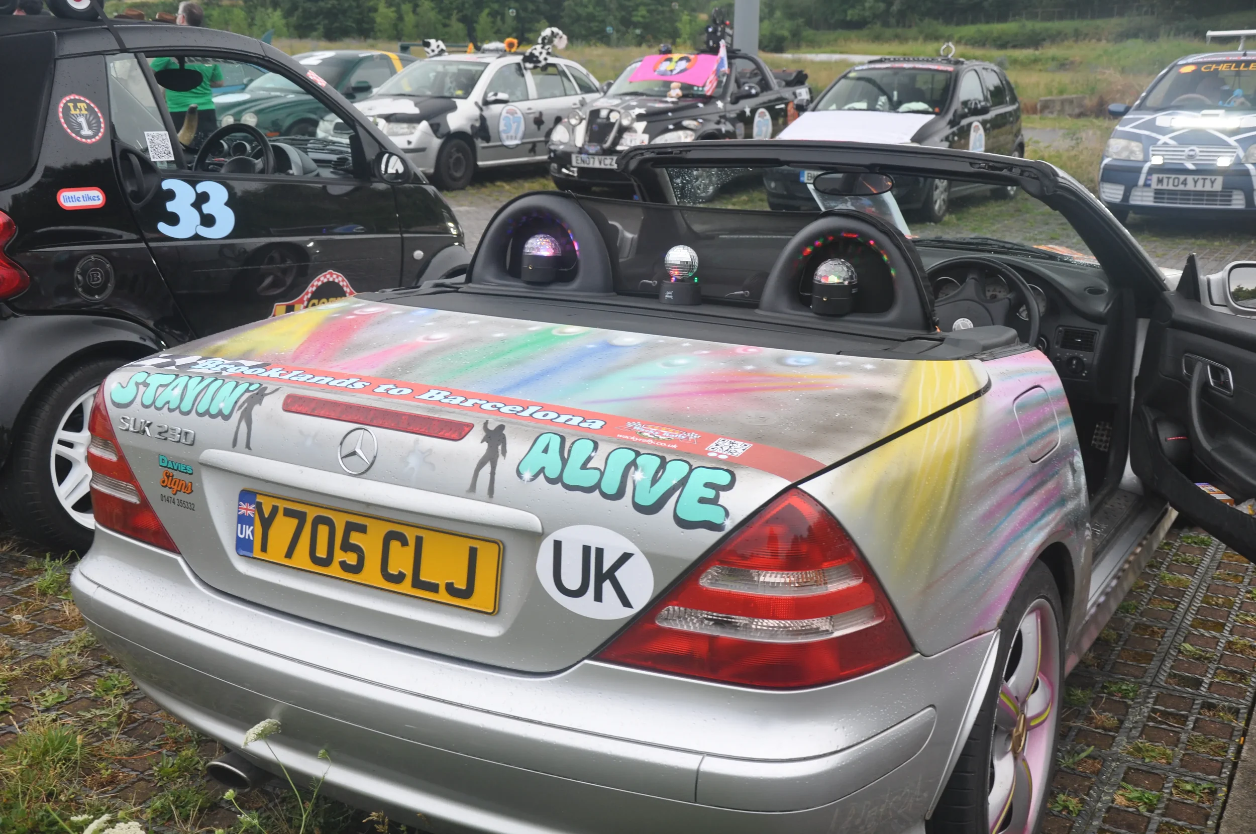 A silver Mercedes convertible decorated with colorful paint splashes and stickers, including phrases such as 'Standing to Barcelona' and 'Alive,' parked among other decorated cars in a parking lot during a rally or event.