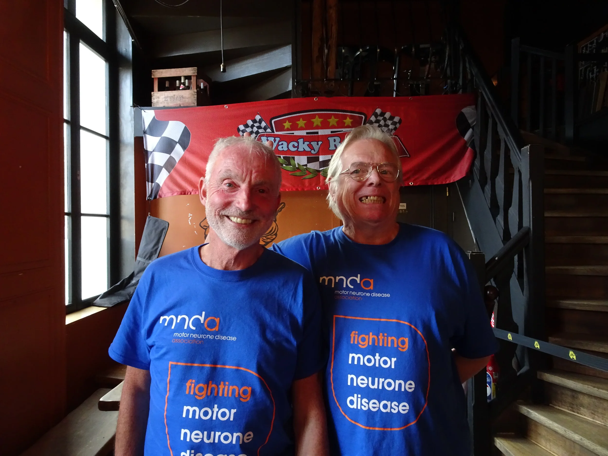 Two smiling older men wearing blue t-shirts that say 'fighting motor neurone disease' standing inside a building near a staircase with a red banner in the background.