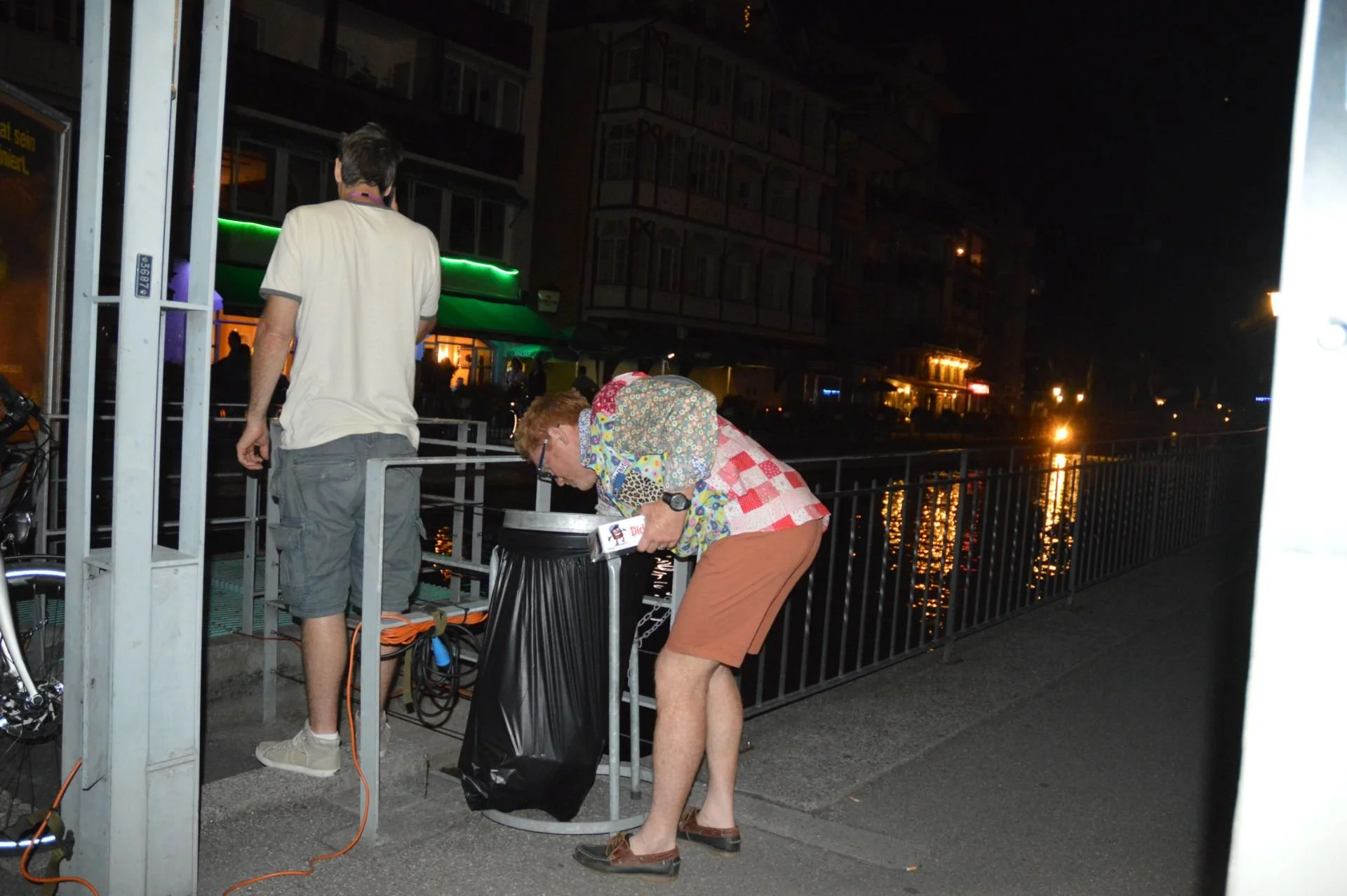 An elderly woman with short red hair, wearing glasses, a colorful floral shirt, and brown shorts, bends over a trash can by the waterfront at night. A younger man with dark hair, wearing a light-colored T-shirt and gray shorts, stands nearby with his