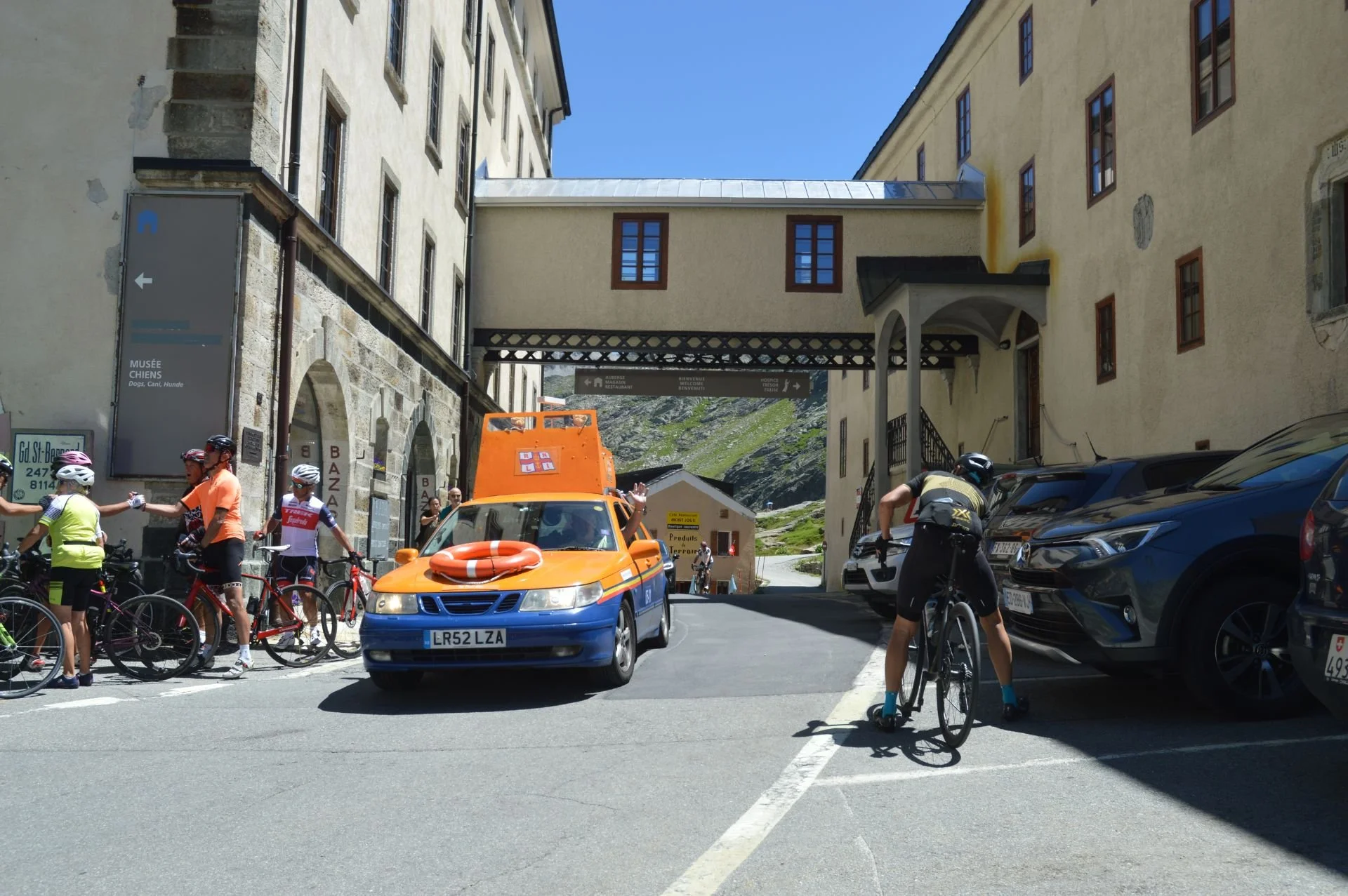 Cyclists and a rescue vehicle in a mountainous town street with historic buildings and a bridge overhead.