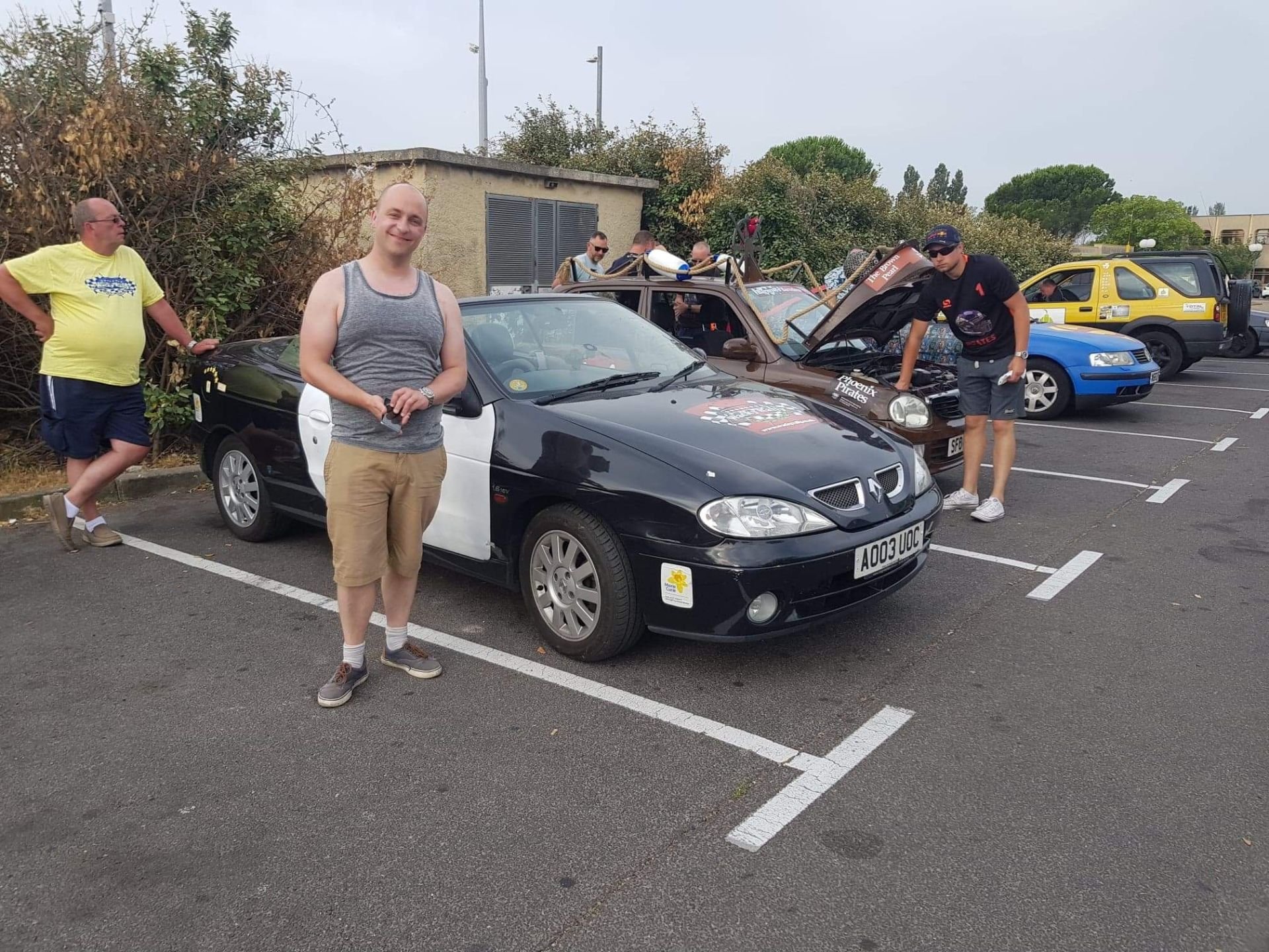 Group of four men unintentionally dressed casually, standing in a parking lot next to various cars, including a black and white convertible with a European license plate and a brown car with an open hood, with trees and a building in the background.