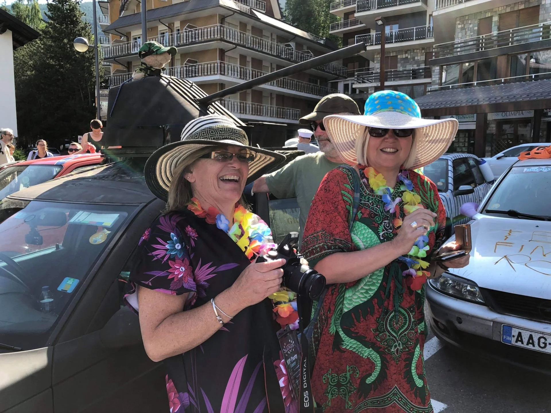 Group of excited women wearing colorful tropical dresses, large sun hats, and leis, standing outdoors near a car, with others and buildings in the background.