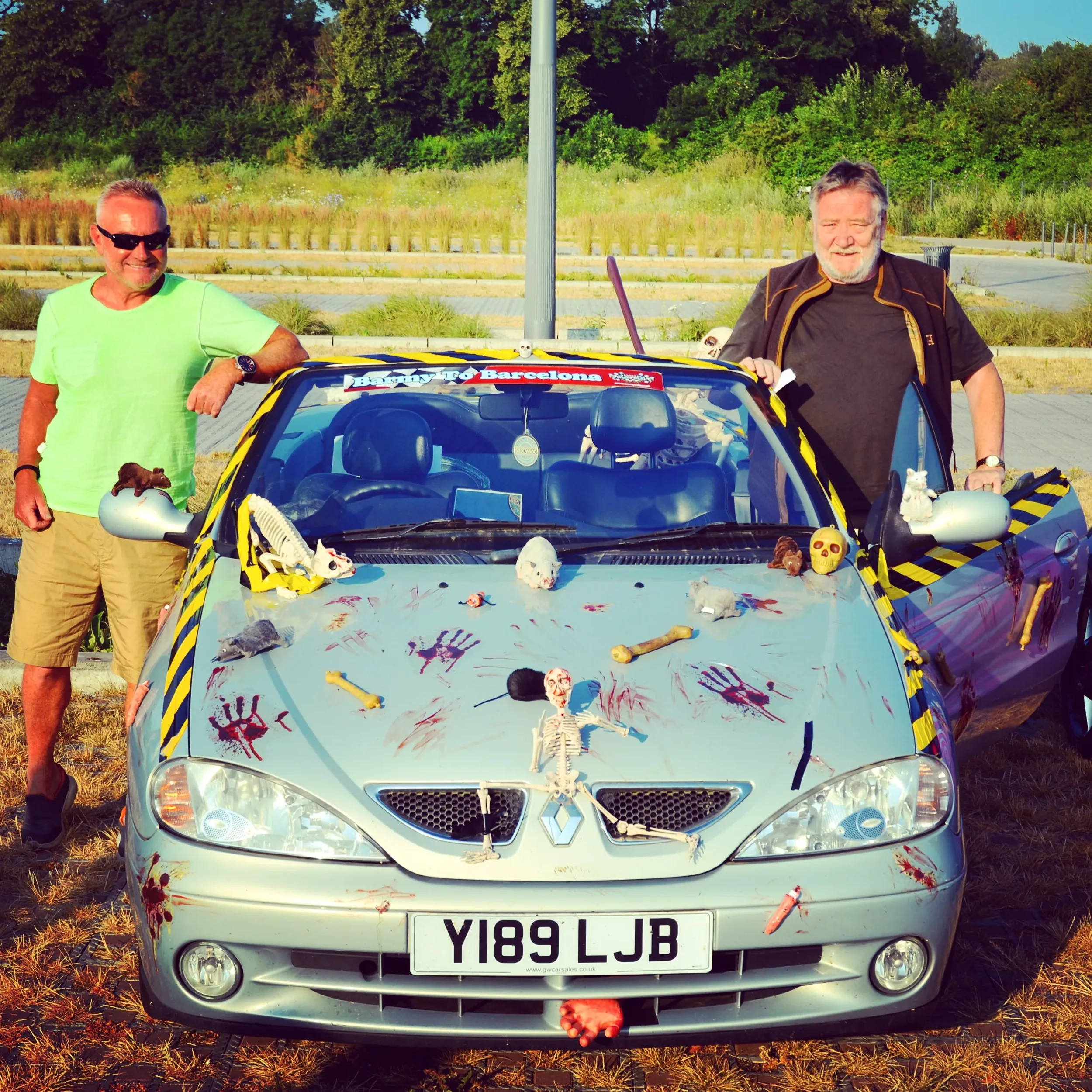 A silver Renault convertible decorated with Halloween props and fake blood, with two men standing beside it. The car has stuffed animals, skeletons, and toy bones on the hood and sides, and a sign that reads 'Journey to Barcelona.' The men are smilin
