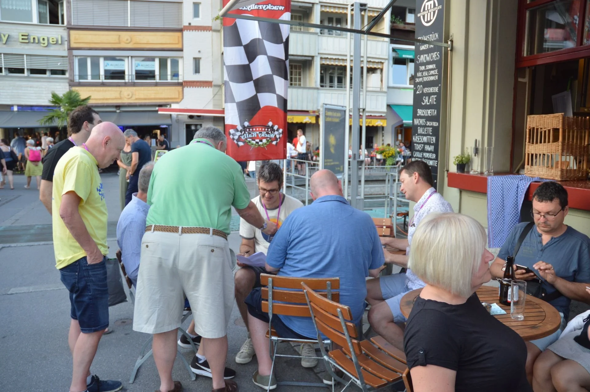 People dining and socializing outdoors at a restaurant or cafe, with some standing and others seated at tables.