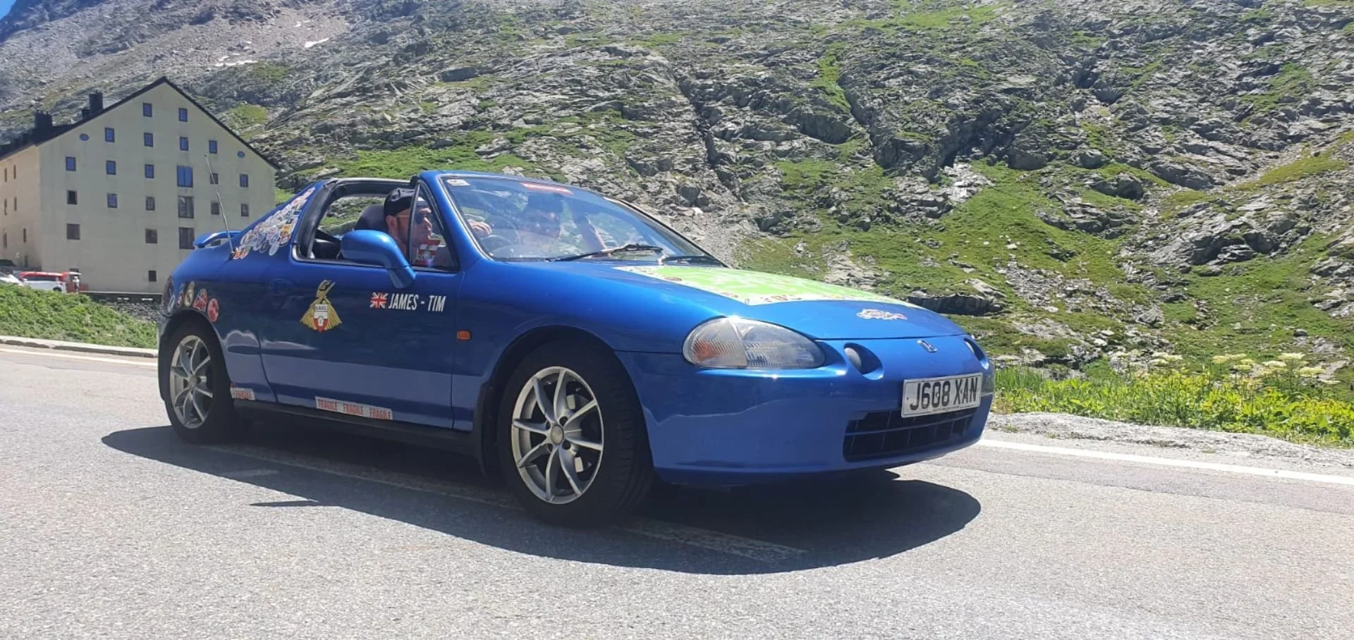 Blue convertible car with racing decals, parked on a mountain road with rocky terrain and green grass in the background.