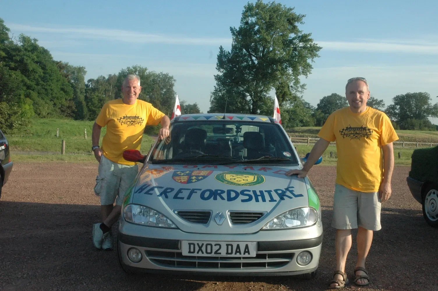 Two men standing next to a silver Renault car with a roof mounted with two small flags. Both men are wearing yellow t-shirts and beige shorts, standing on either side of the car in an outdoor parking lot with a grassy field and trees in the backgroun