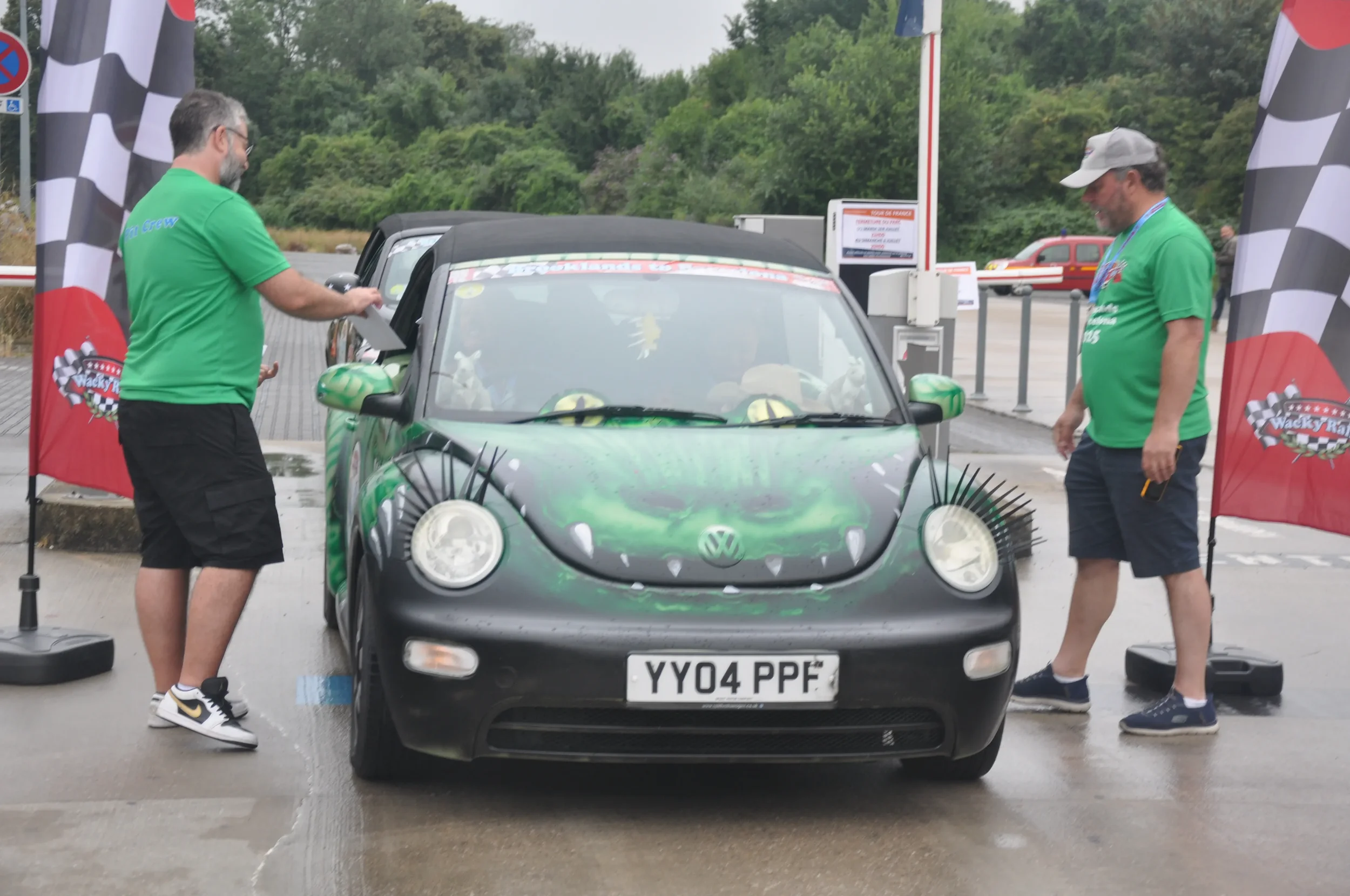 A Volkswagen car with a custom green dragon-themed paint job, decorated with plush toys and spikes. Two men in green shirts are standing beside it, one on each side, at a race event with checkered flags and banners in the background.