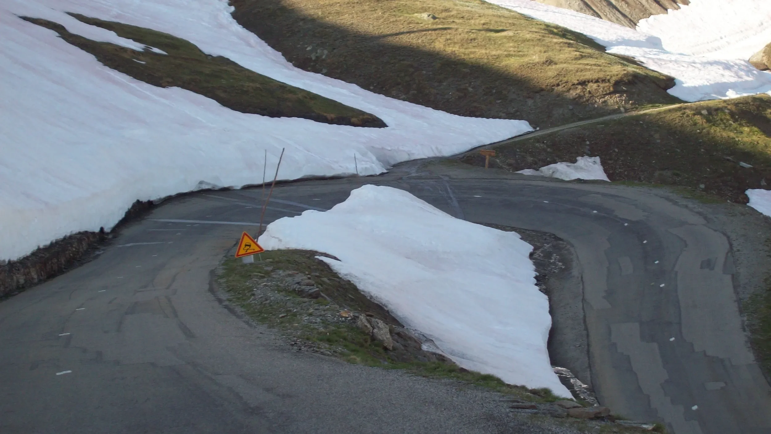 A winding mountain road with patches of snow on the sides and a slippery road warning sign.