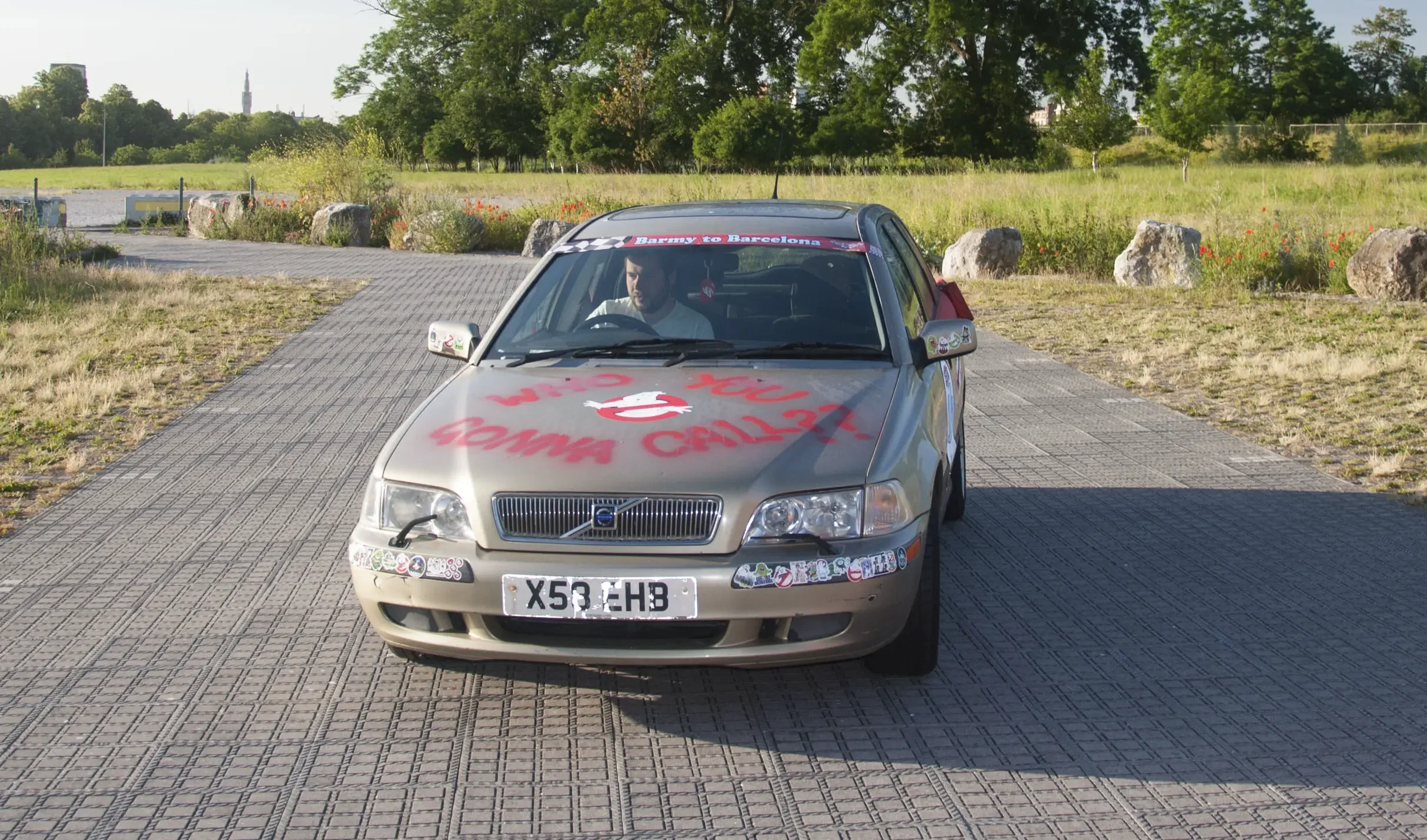A silver Volvo car with bumper stickers and red and pink writing on the hood, parked on a paved pathway in a park-like area with green trees, rocks, and grass in the background.