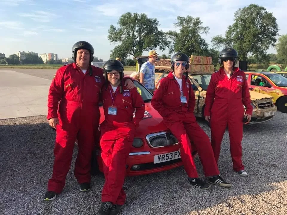 Four women wearing red racing suits and helmets pose in front of a small red car, standing on a gravel surface with a few other cars and a man in a hat in the background, outdoors under a partly cloudy sky.