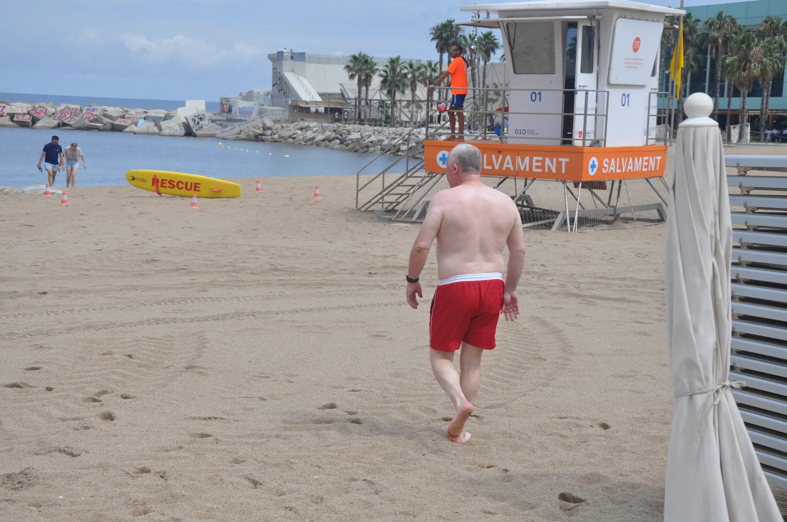 A man in red shorts walking on a sandy beach near a lifeguard tower with rescue equipment and a kayak, with two people walking by the water in the background.