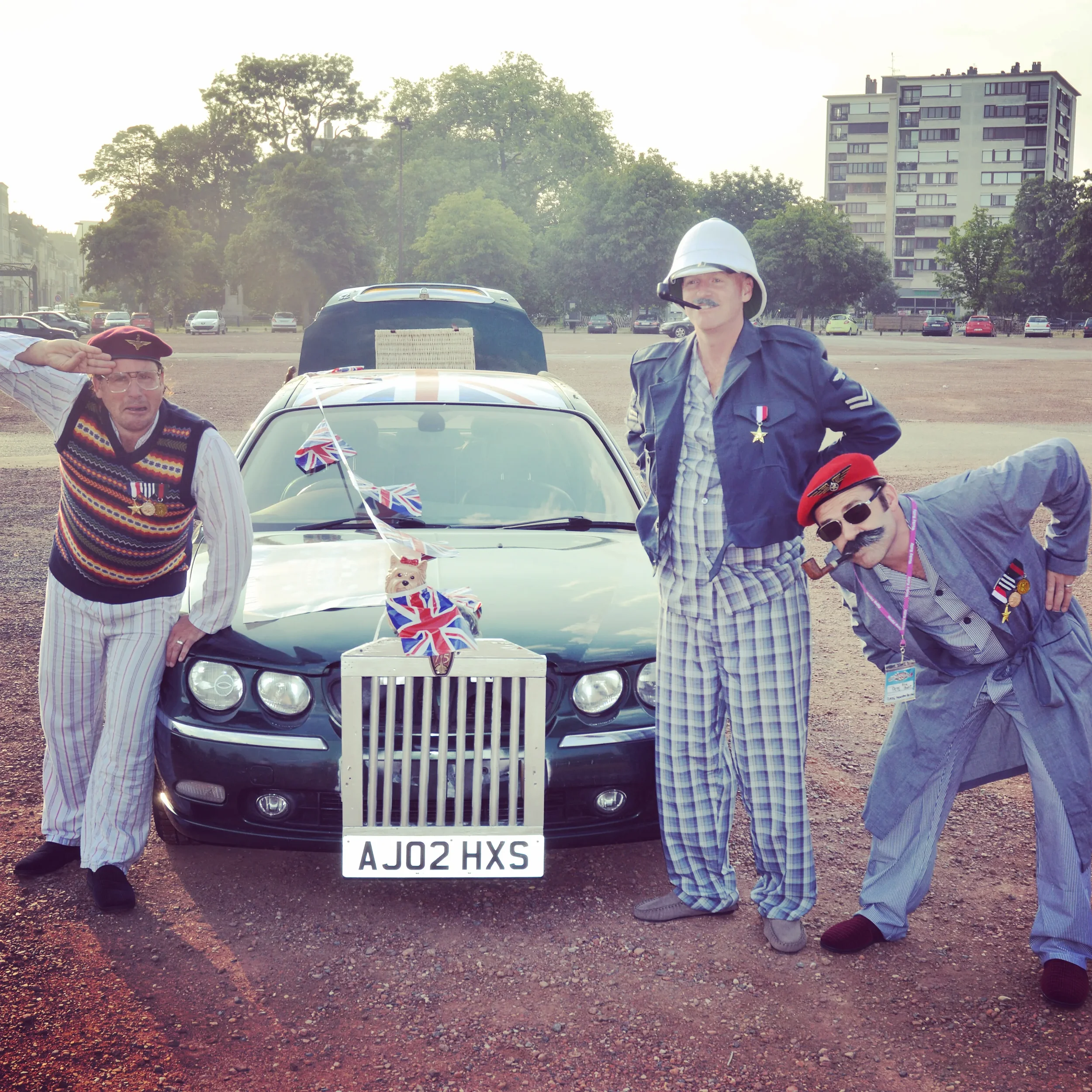 People dressed in vintage British soldier costumes standing in front of a black car decorated with Union Jack flags, in an open outdoor area with trees and buildings in the background.