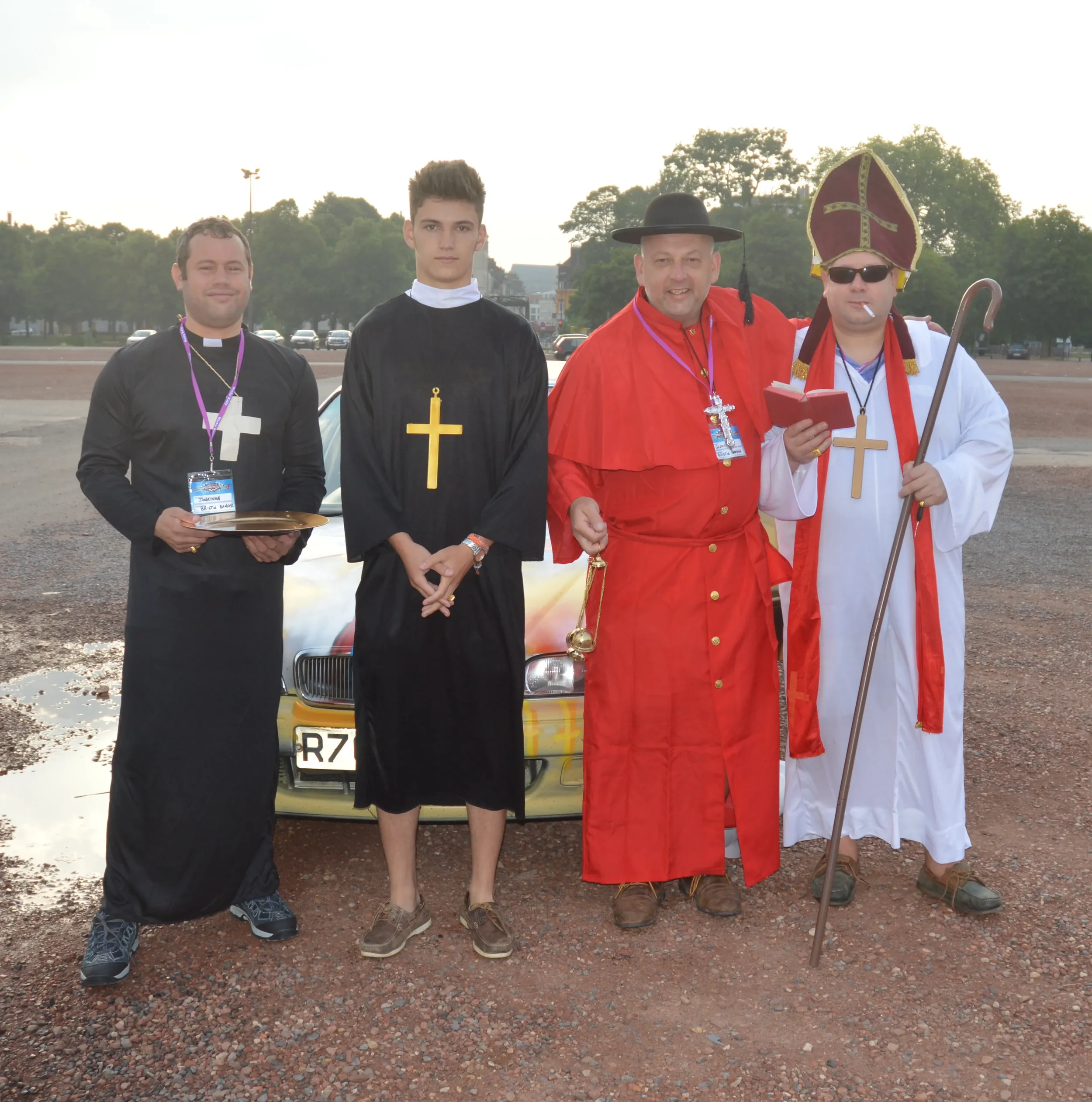 Four men in religious costumes standing outdoors in front of a yellow car on a gravel surface with trees in the background. One man is dressed as a priest, another as a clergyman in black, one as a cardinal in red, and the fourth as a bishop in white