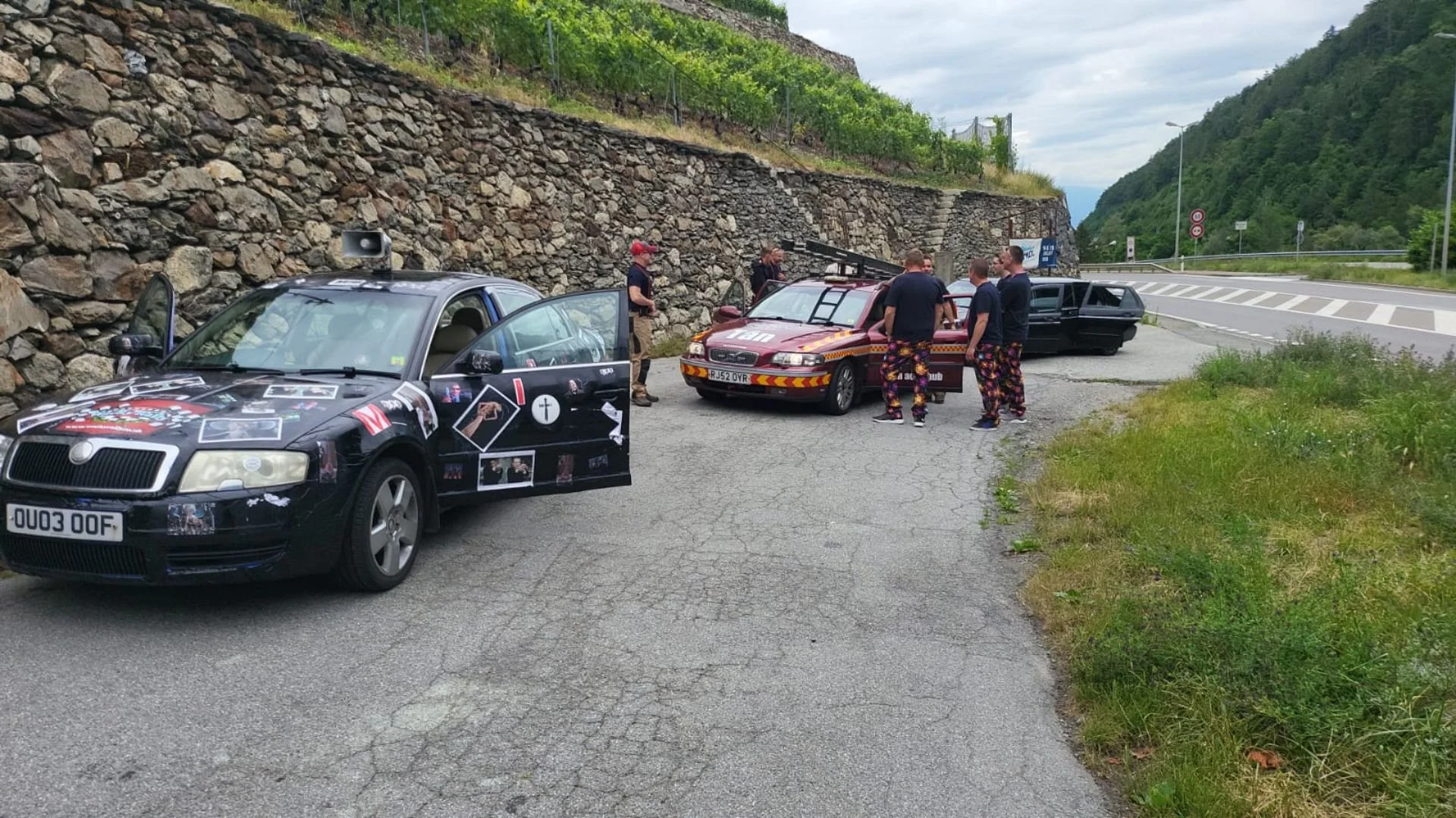 Multiple emergency service vehicles, including a black and a red car, are parked on the side of a mountain road with four men gathered around. The vehicles include a black car with graphics and a red car with reflective markings. The men are wearing 