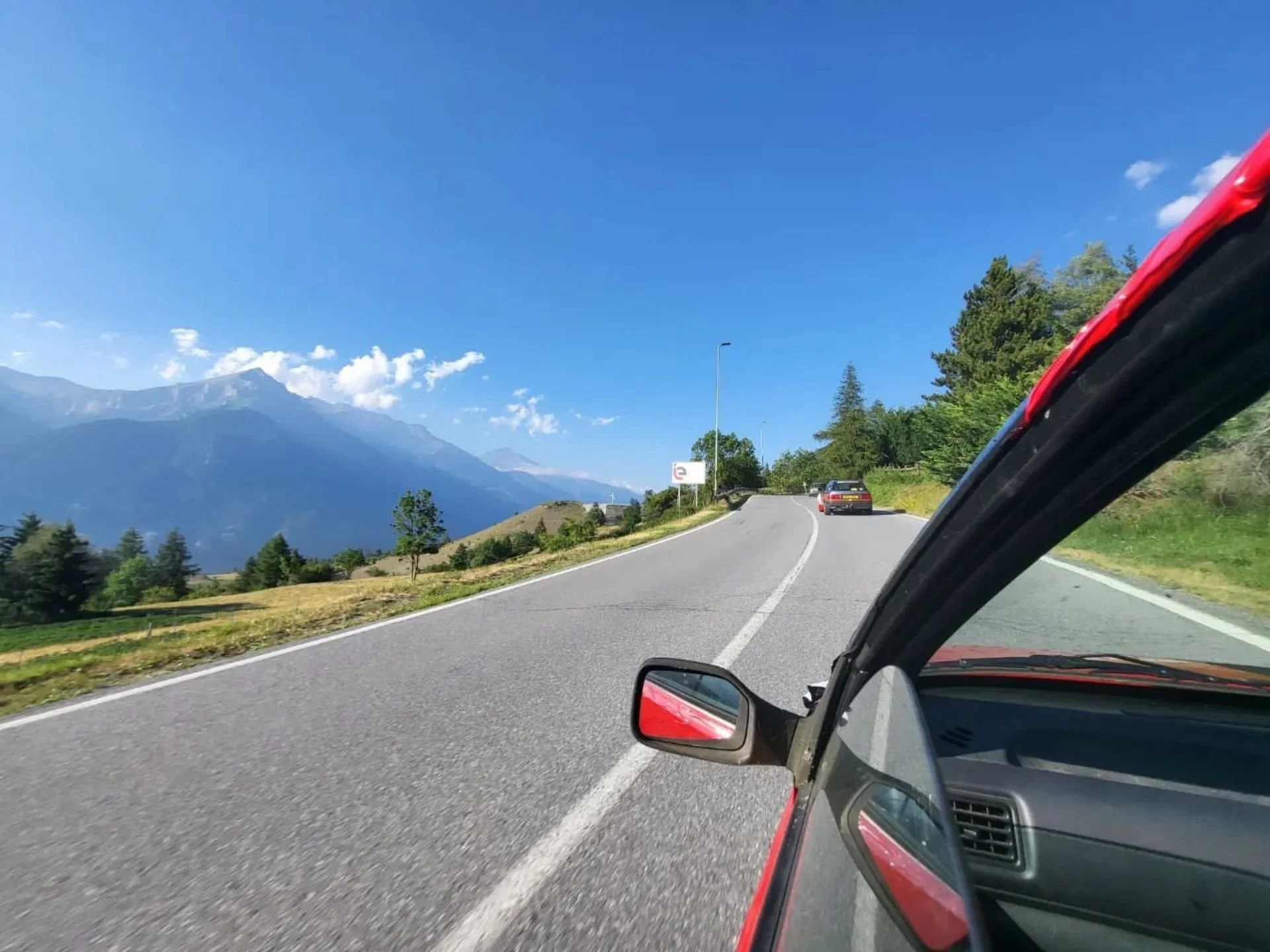 View of a mountain road with cars, trees, mountains, and a blue sky with some clouds.