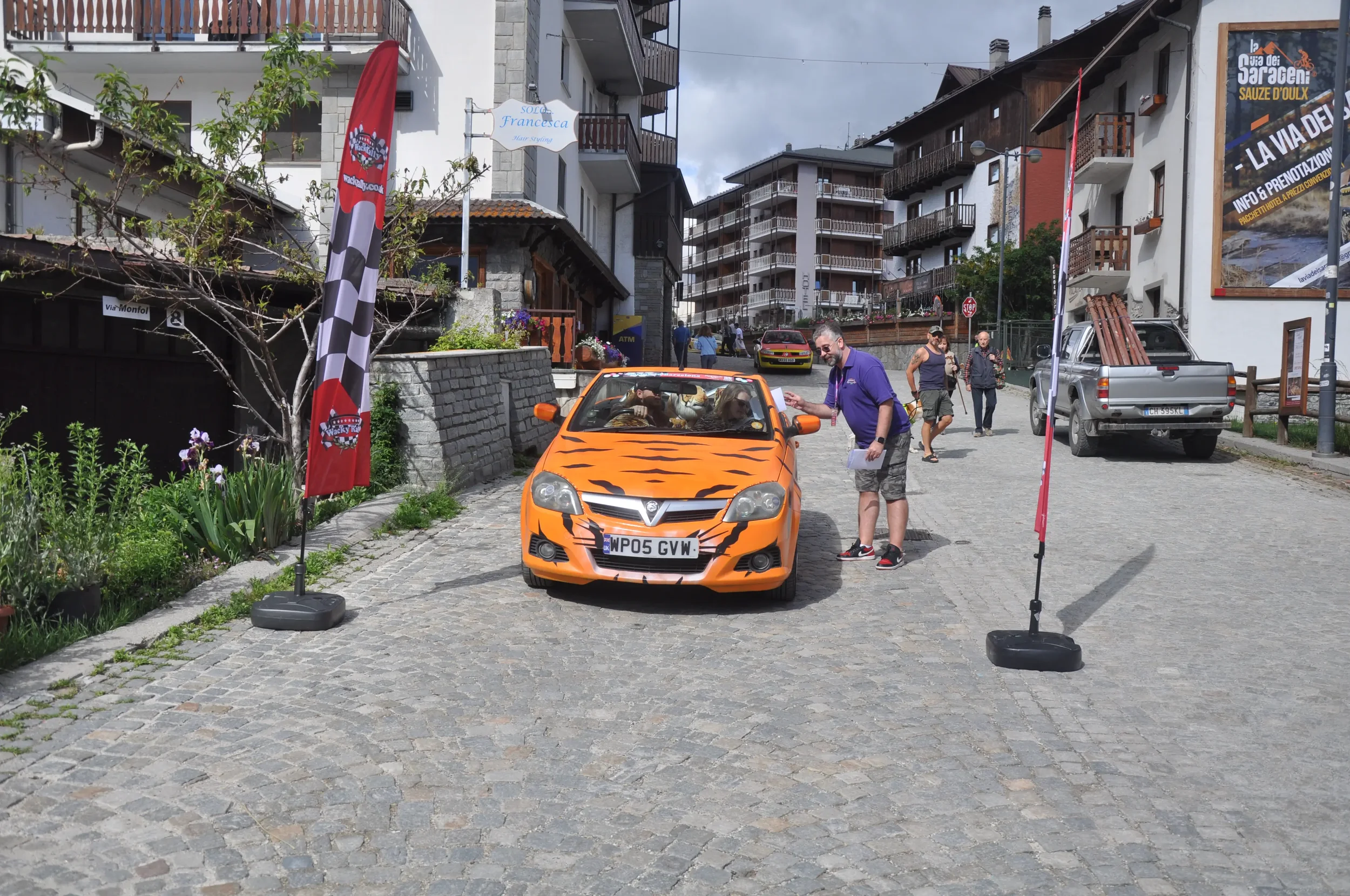A street scene with a small orange car painted with black tiger stripes, parked between two flags on stands. A man in a purple shirt and shorts is talking to or inspecting the car, while two children sit inside. Several people are walking on the cobb