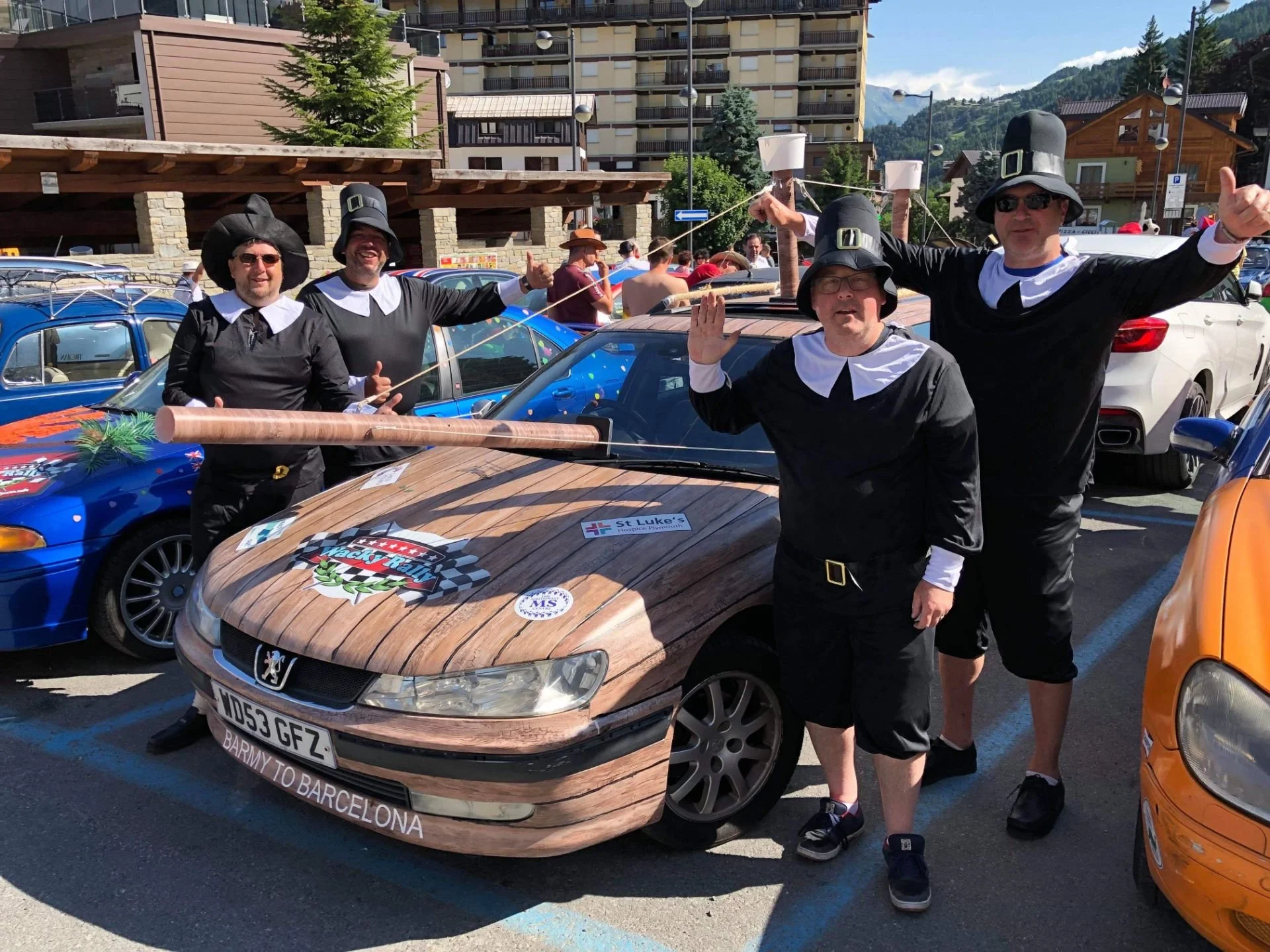 Four men dressed in costumes resembling Pilgrims from Thanksgiving standing next to a car decorated with a wooden plank design, at an outdoor car event.