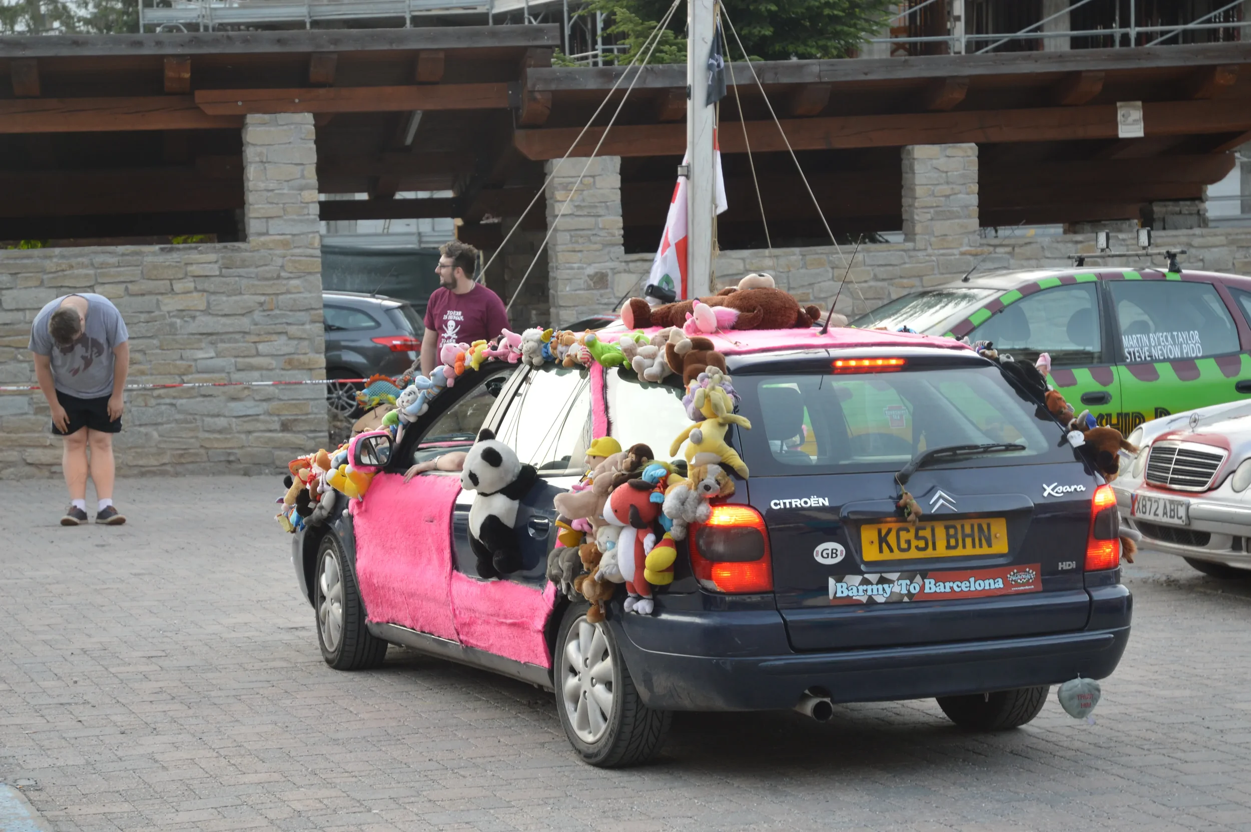 A decorated car with many plush toys attached, parked in a lot near a building with stone walls. Two men are nearby, one bowing, the other standing and smiling.