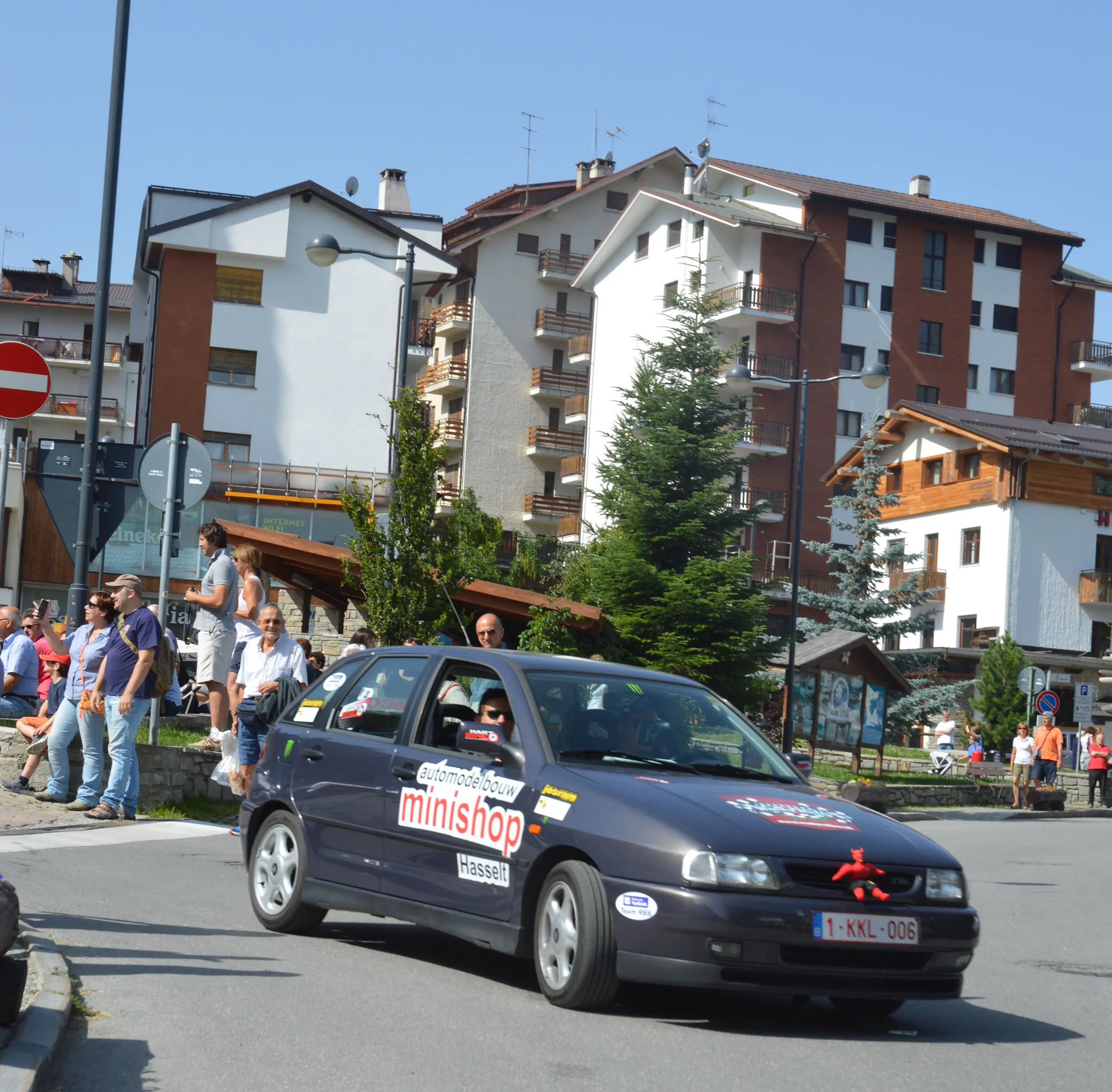 A black hatchback car decorated with rally stickers, including 'minishop' and 'auto mobil bouw', driving on a street in a mountain town with apartment buildings and people walking and sitting nearby.